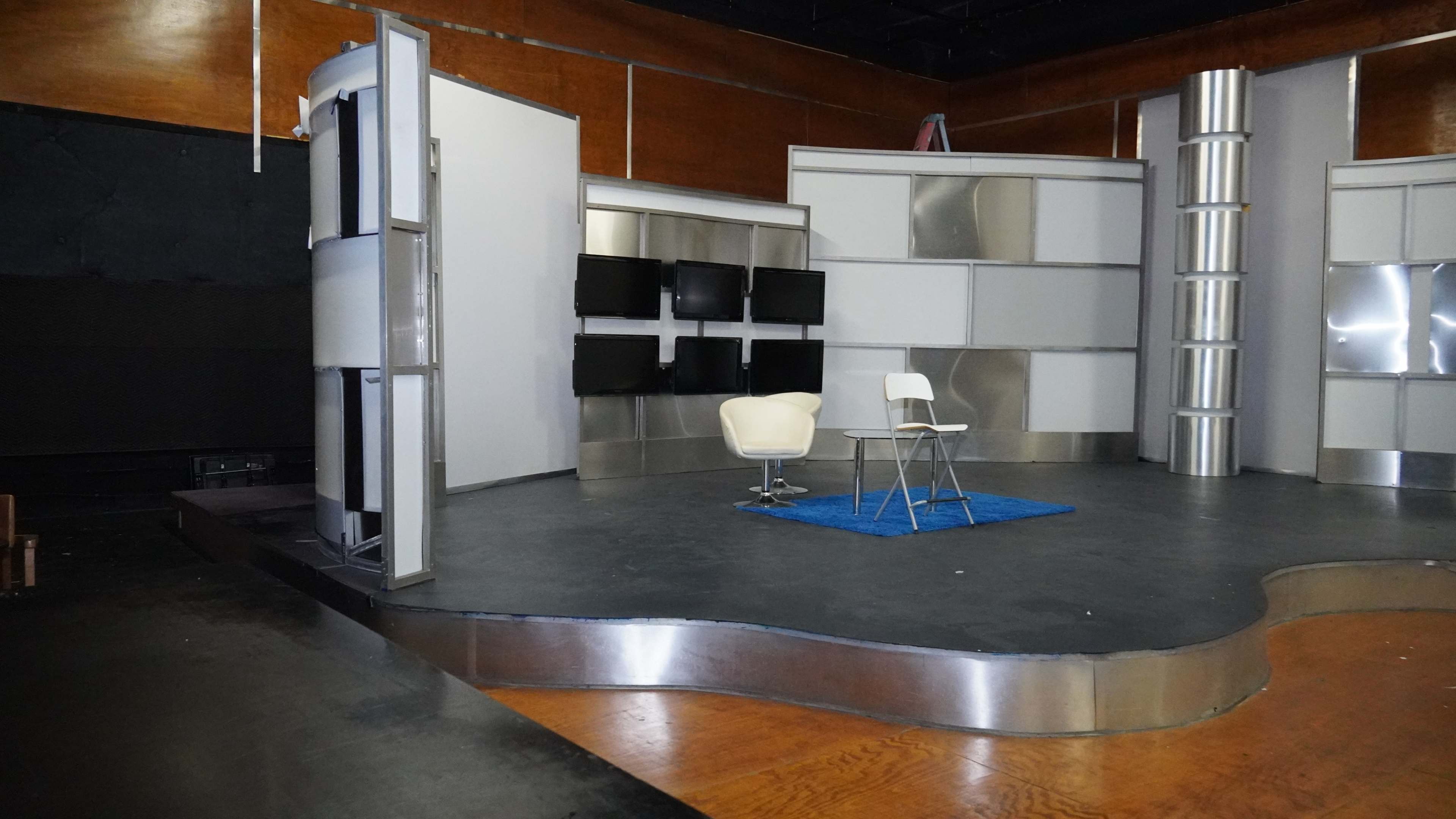 The image shows an empty television studio set featuring a chair and a small table on a curved stage with metallic screens in the background.