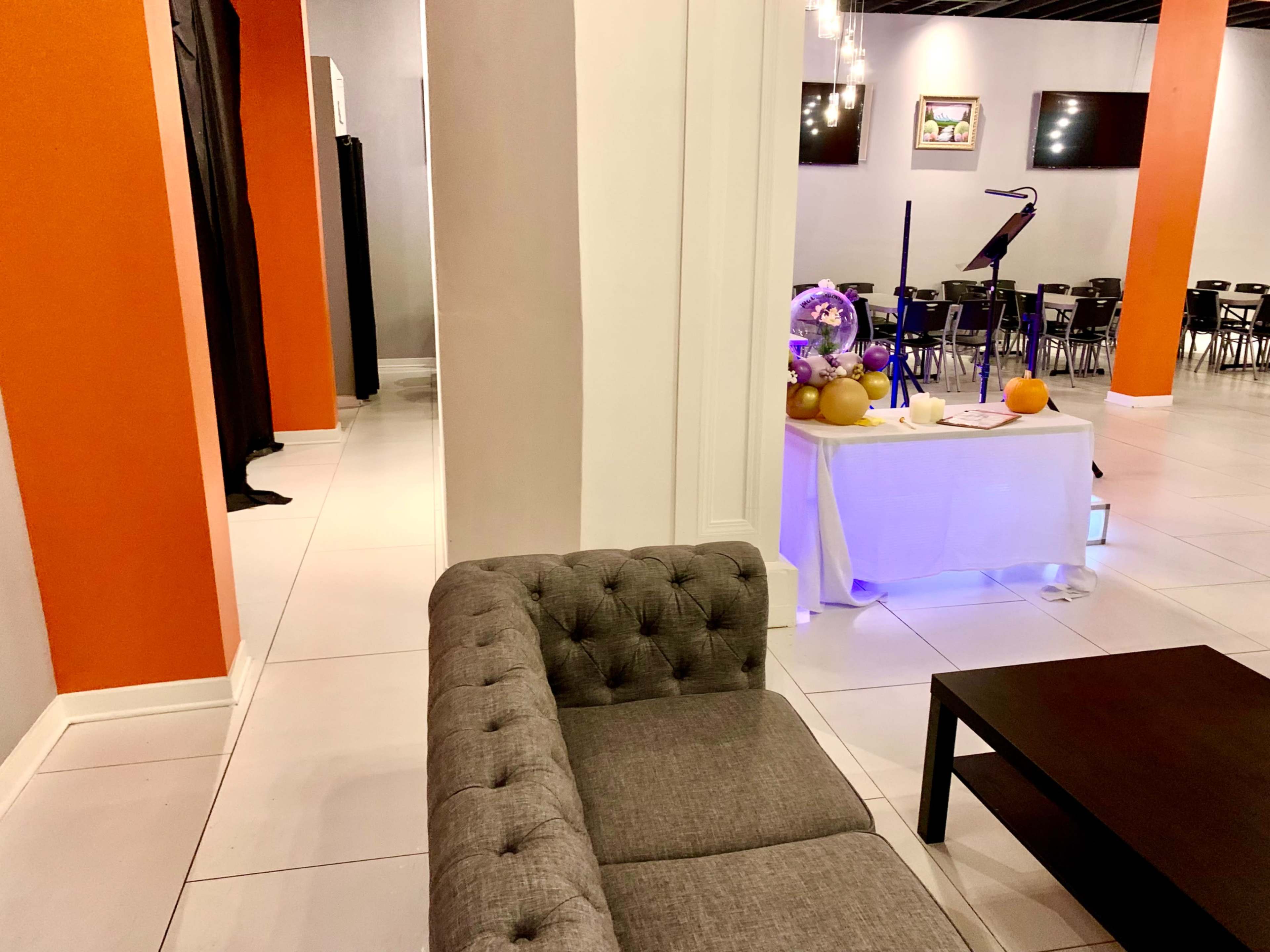 The image shows a spacious indoor venue with a gray tufted couch on the left, a black curtain on the left side, and tables set up in the background for an event.