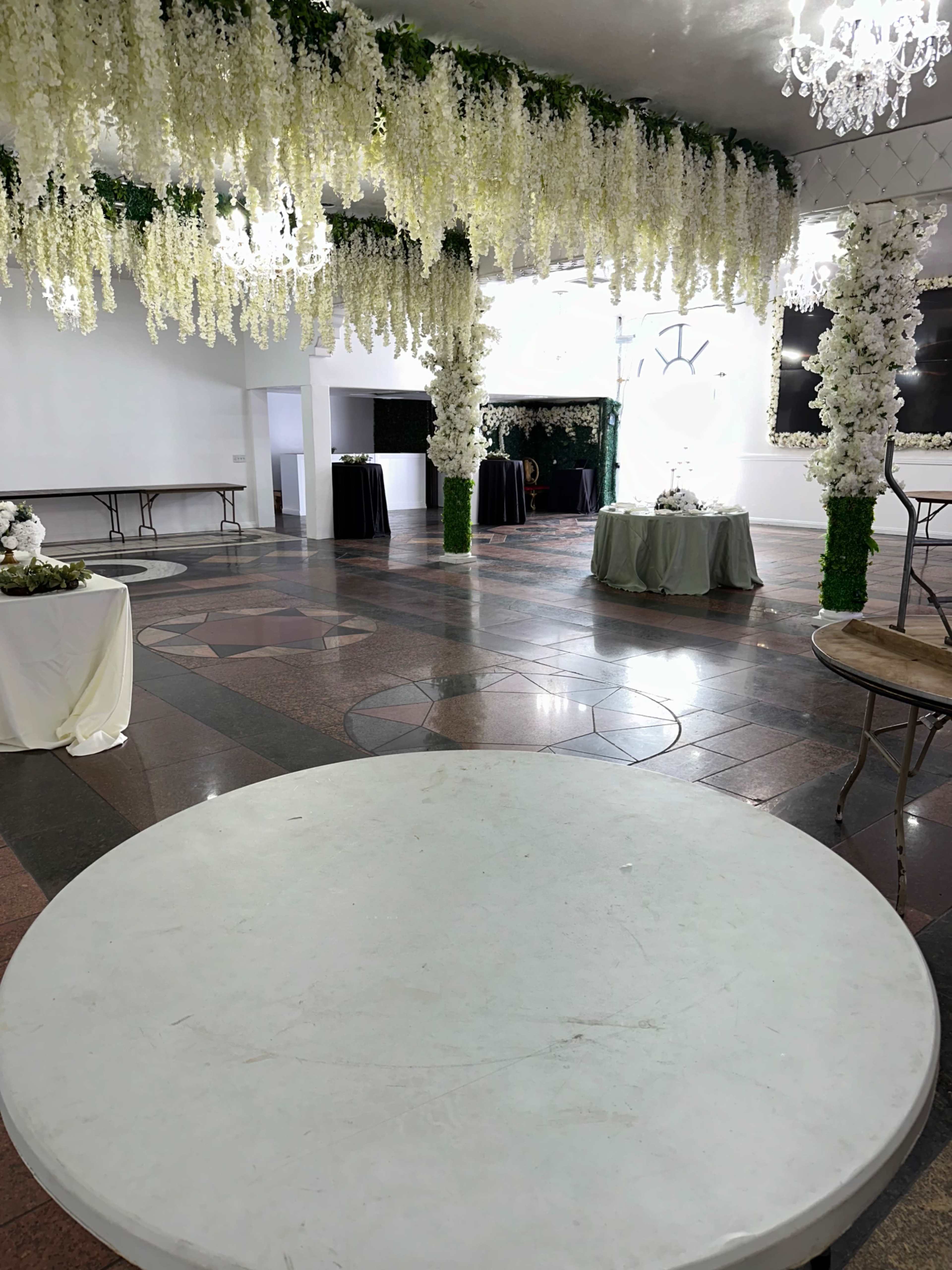 The image shows a large, elegantly decorated event space with hanging floral arrangements, round tables, and a polished floor.