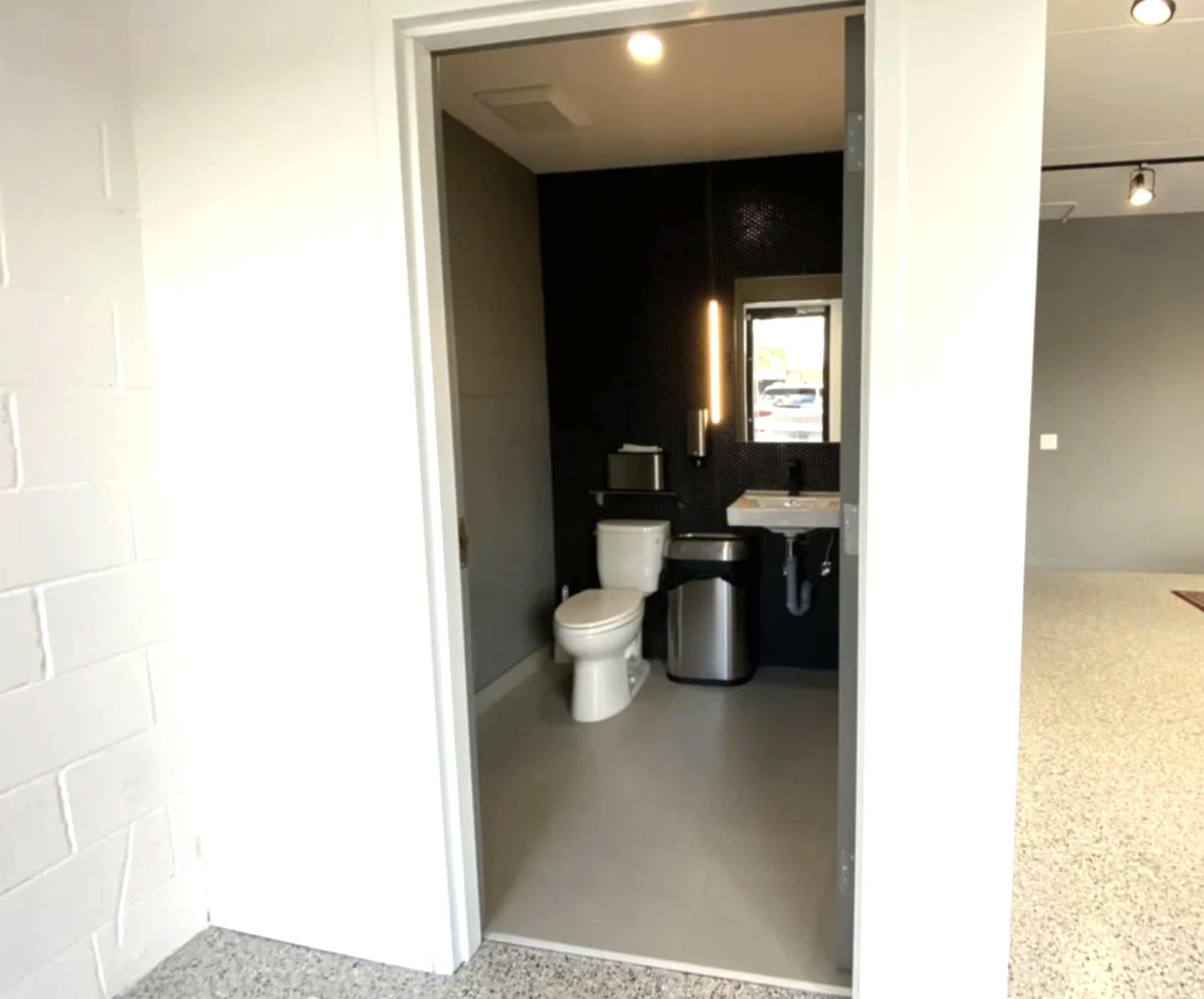 The image shows a clean restroom with a toilet, sink, and a waste bin, viewed from an open doorway.