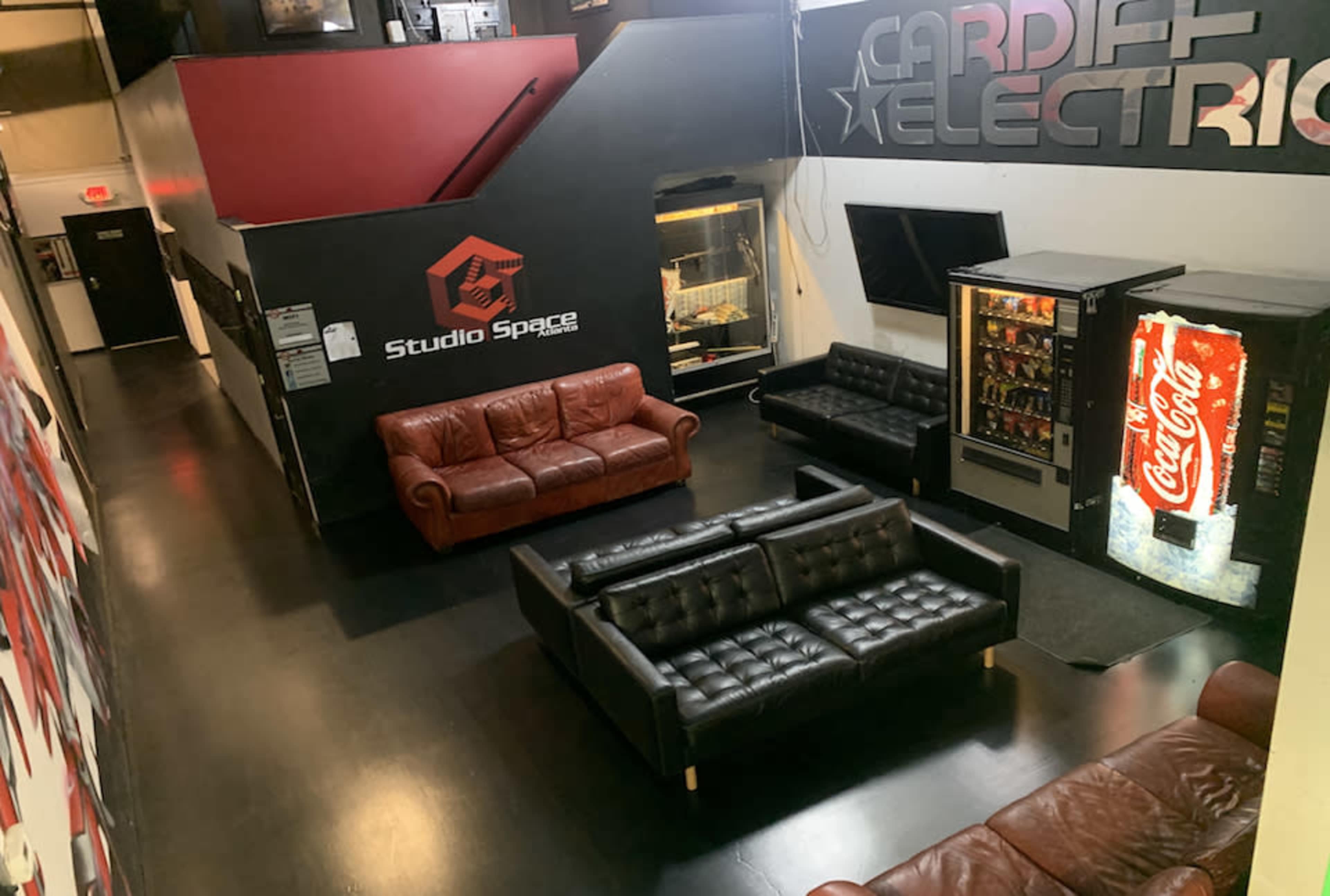 The image shows a spacious lounge area with two leather sofas, a vending machine, and a wall featuring logos, including one for "Studio Space."