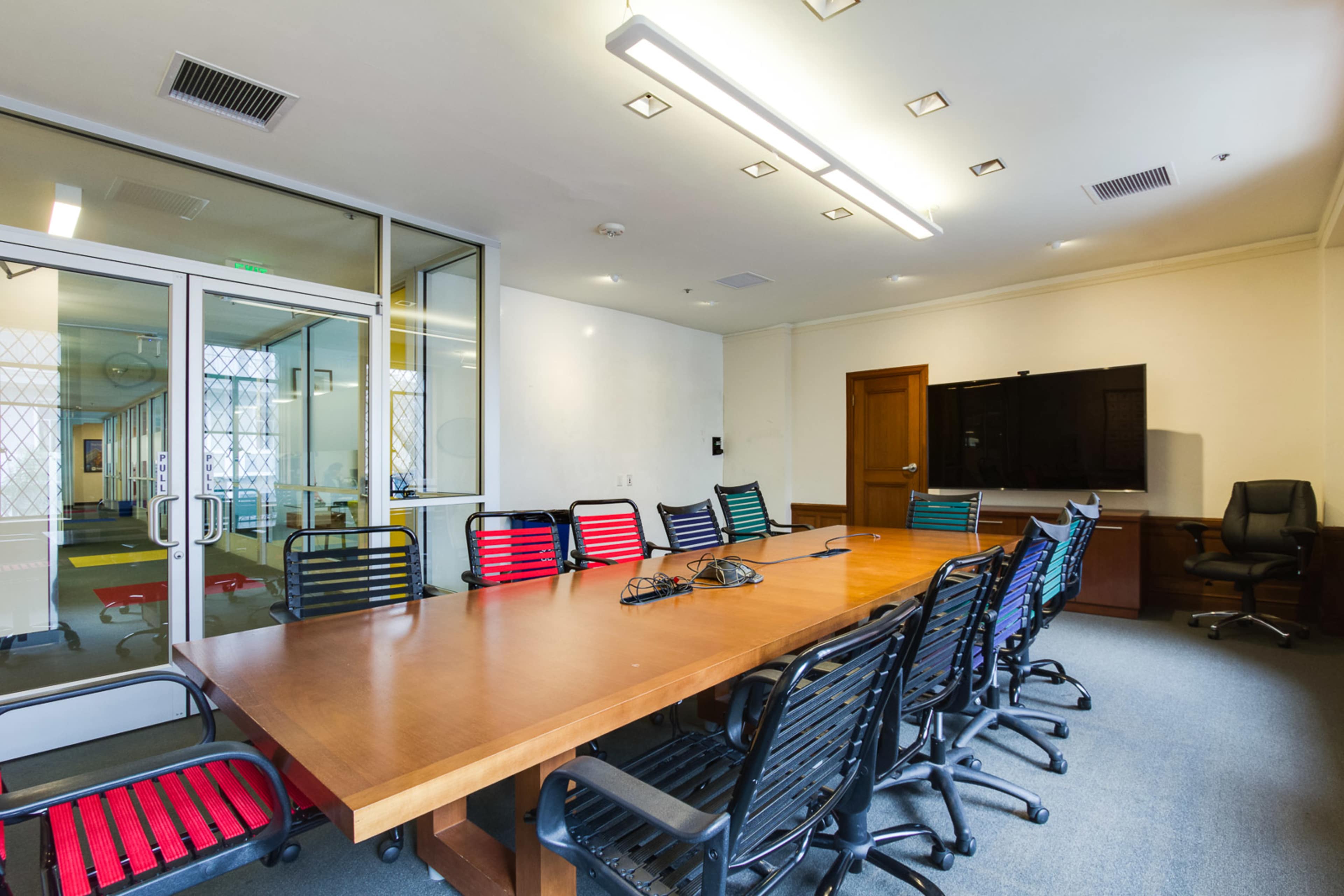 A modern conference room features a large wooden table surrounded by colorful chairs, with a television mounted on the wall and glass doors leading to another space.
