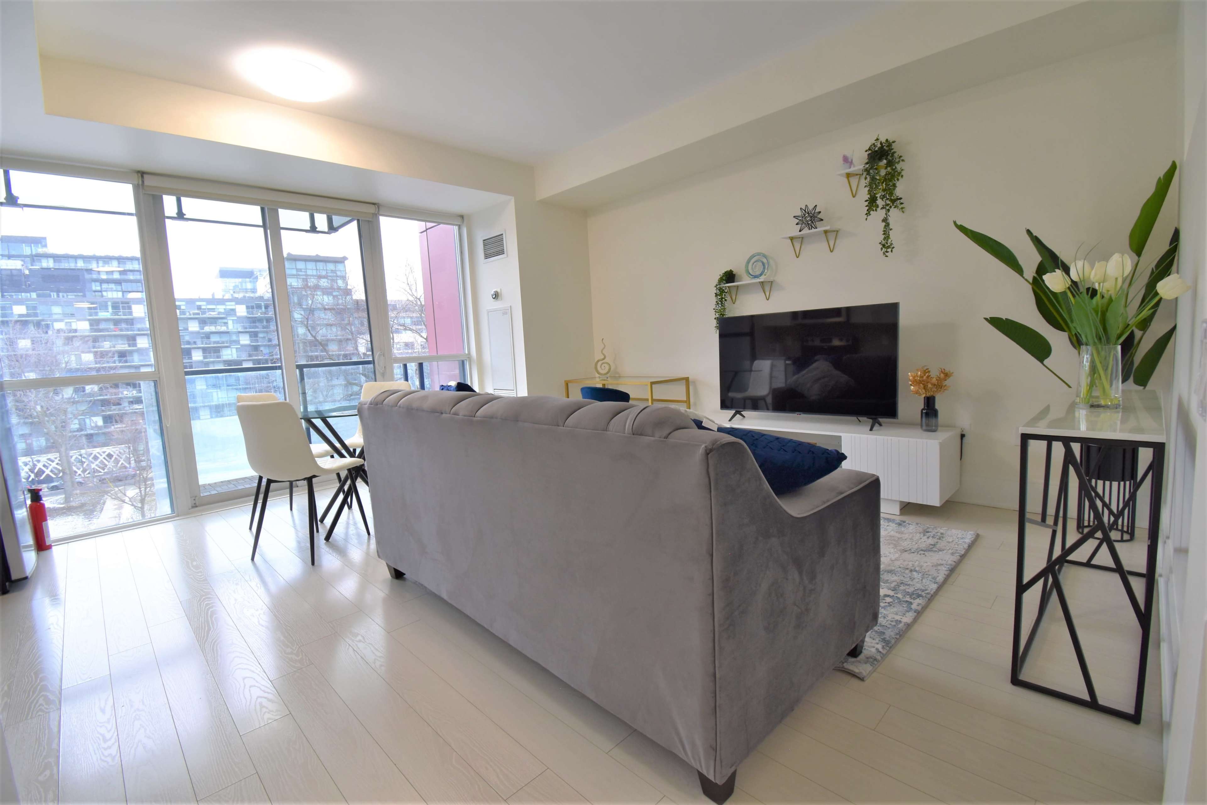 The image shows a modern living room with a gray sofa, a television on a white media unit, and a large window allowing natural light to enter.