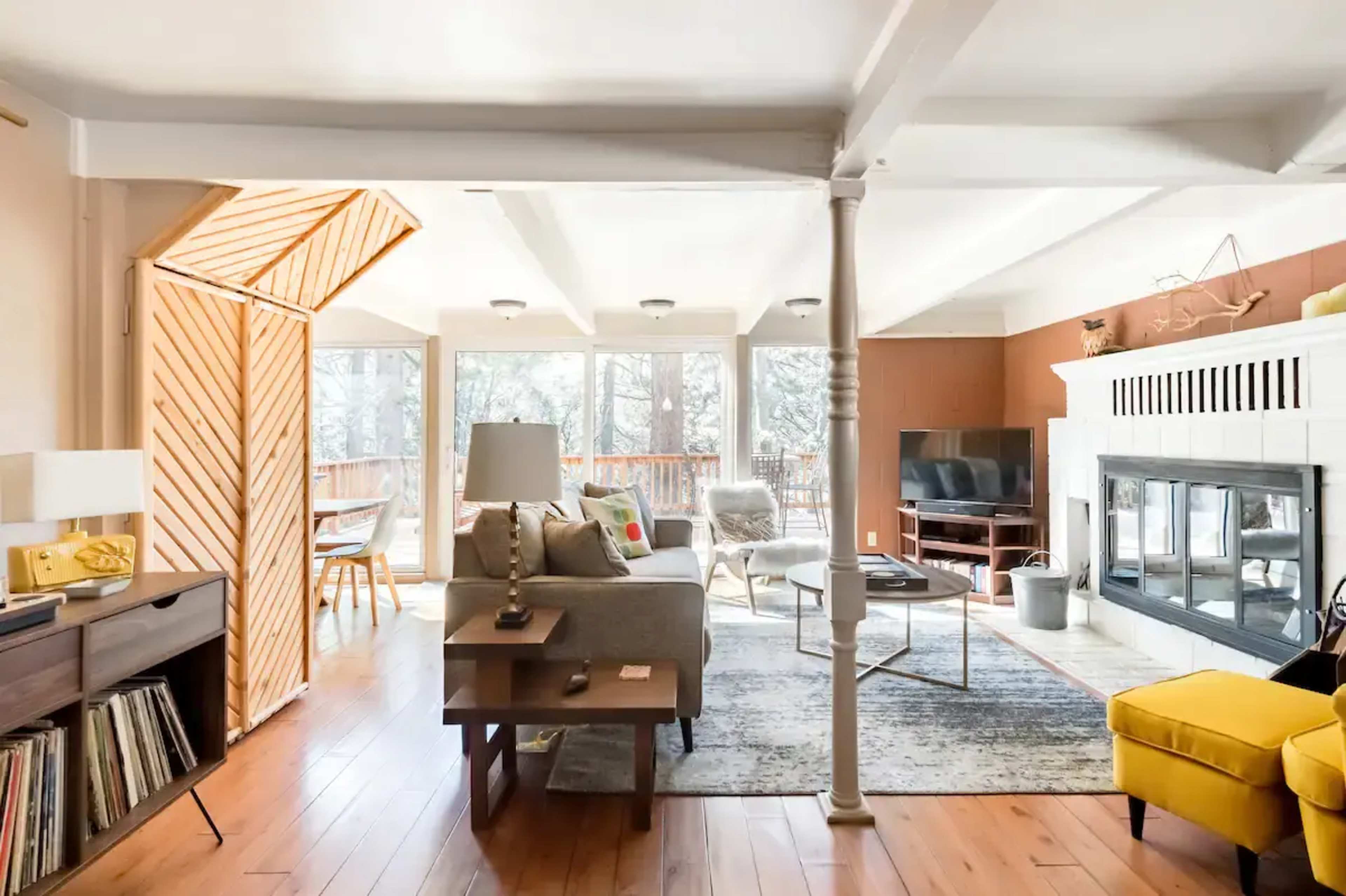 The living room features a neutral-colored sofa, wooden flooring, large windows, and a fireplace, creating an open and airy atmosphere.