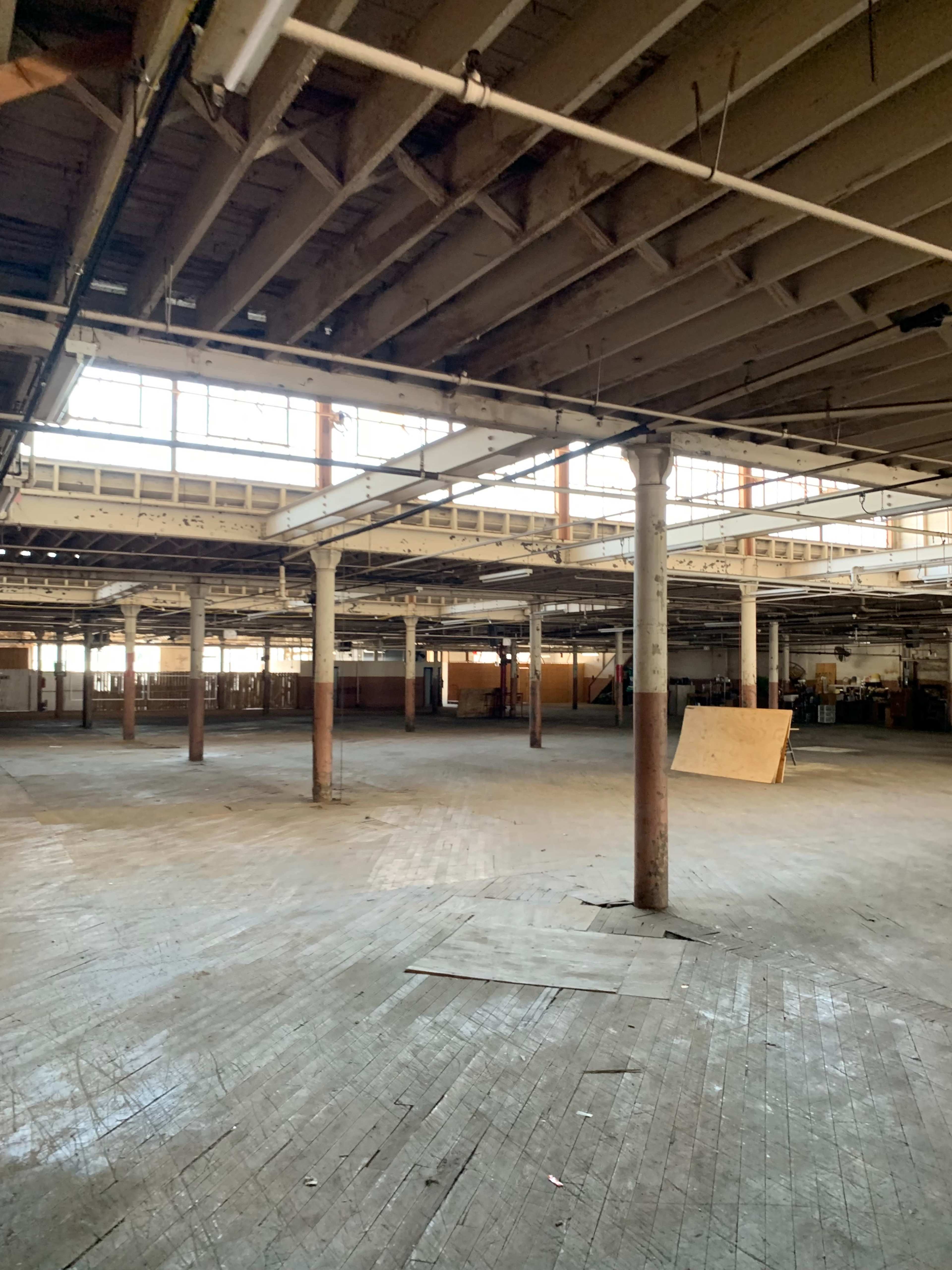 The image shows an empty warehouse interior with exposed beams, wooden floors, and large windows allowing natural light to enter.
