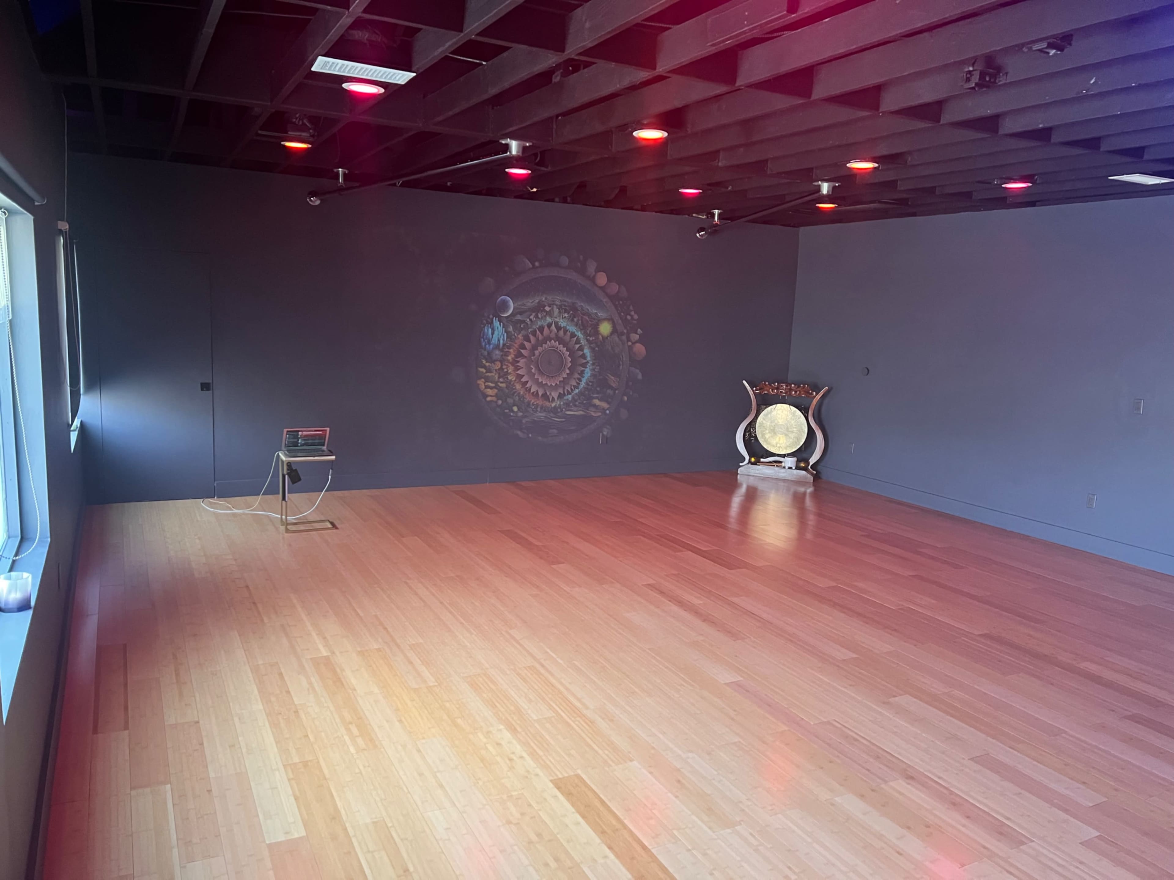 The image shows a spacious, empty room with wooden flooring, a projector display on one wall, and a decorative gong positioned in the corner.