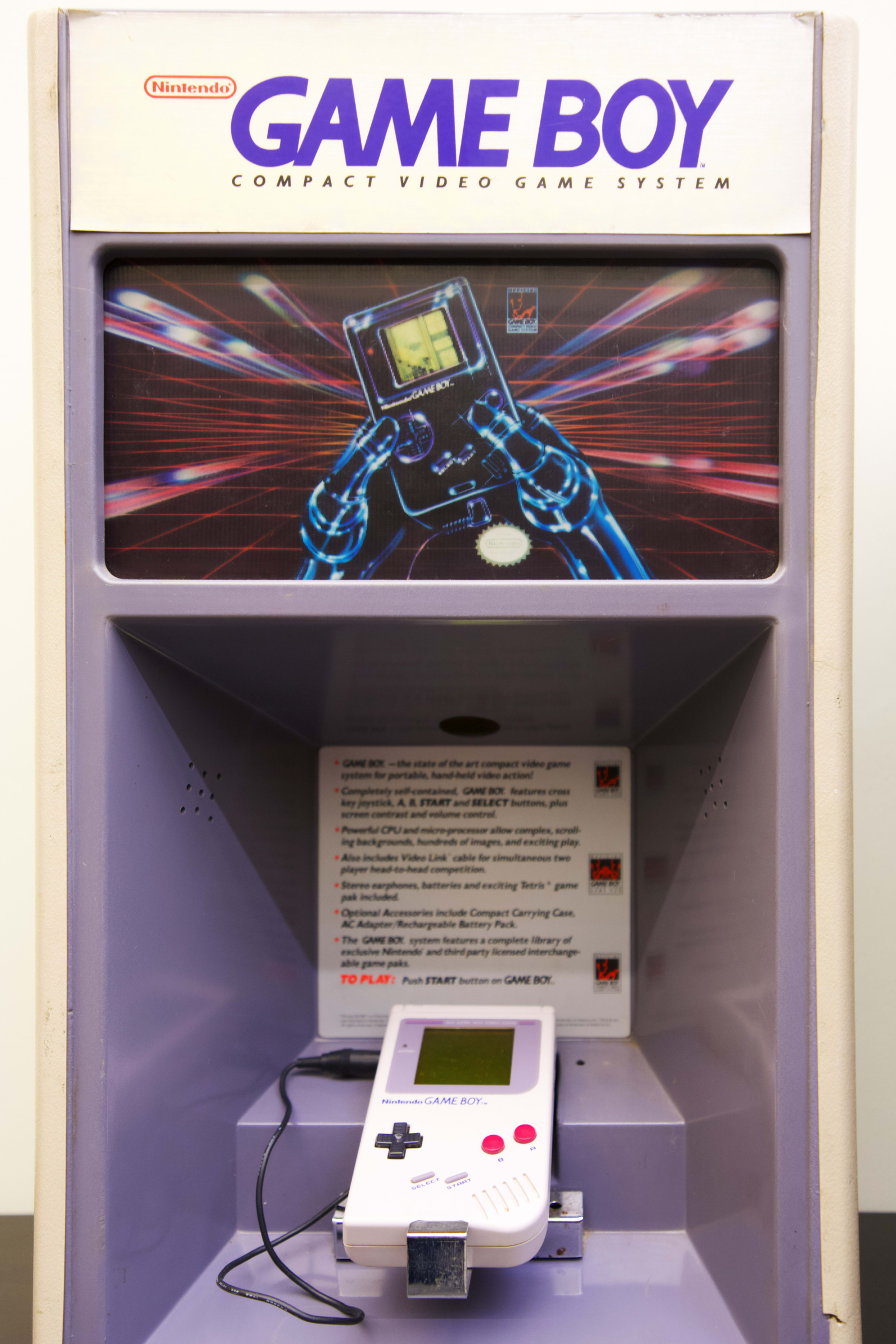 A Nintendo Game Boy display unit featuring a white Game Boy console connected to a stand, with a colorful graphic backdrop.