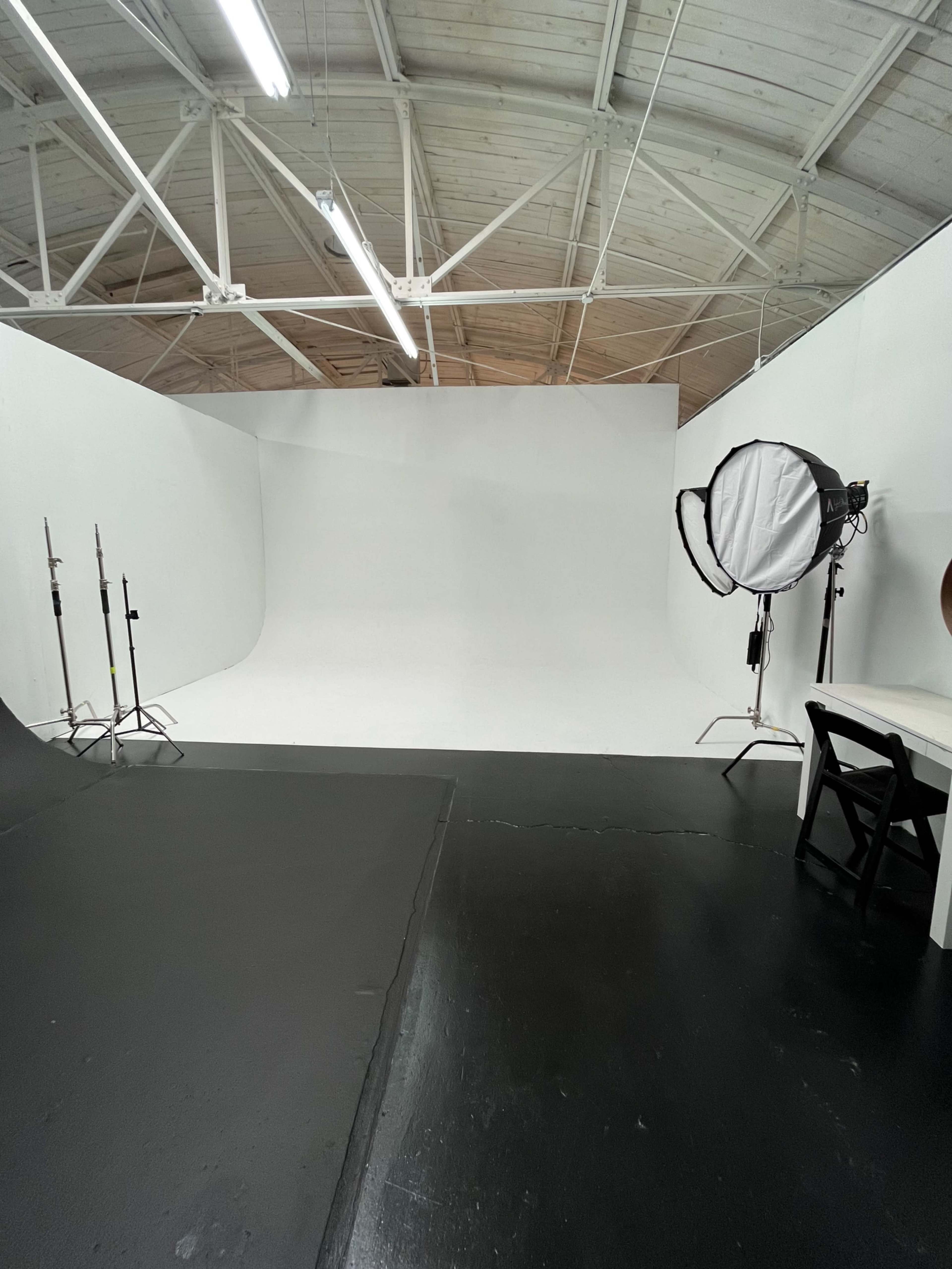 The image shows a photography studio featuring a white backdrop, lighting equipment, and a table with a chair in a spacious, empty area.