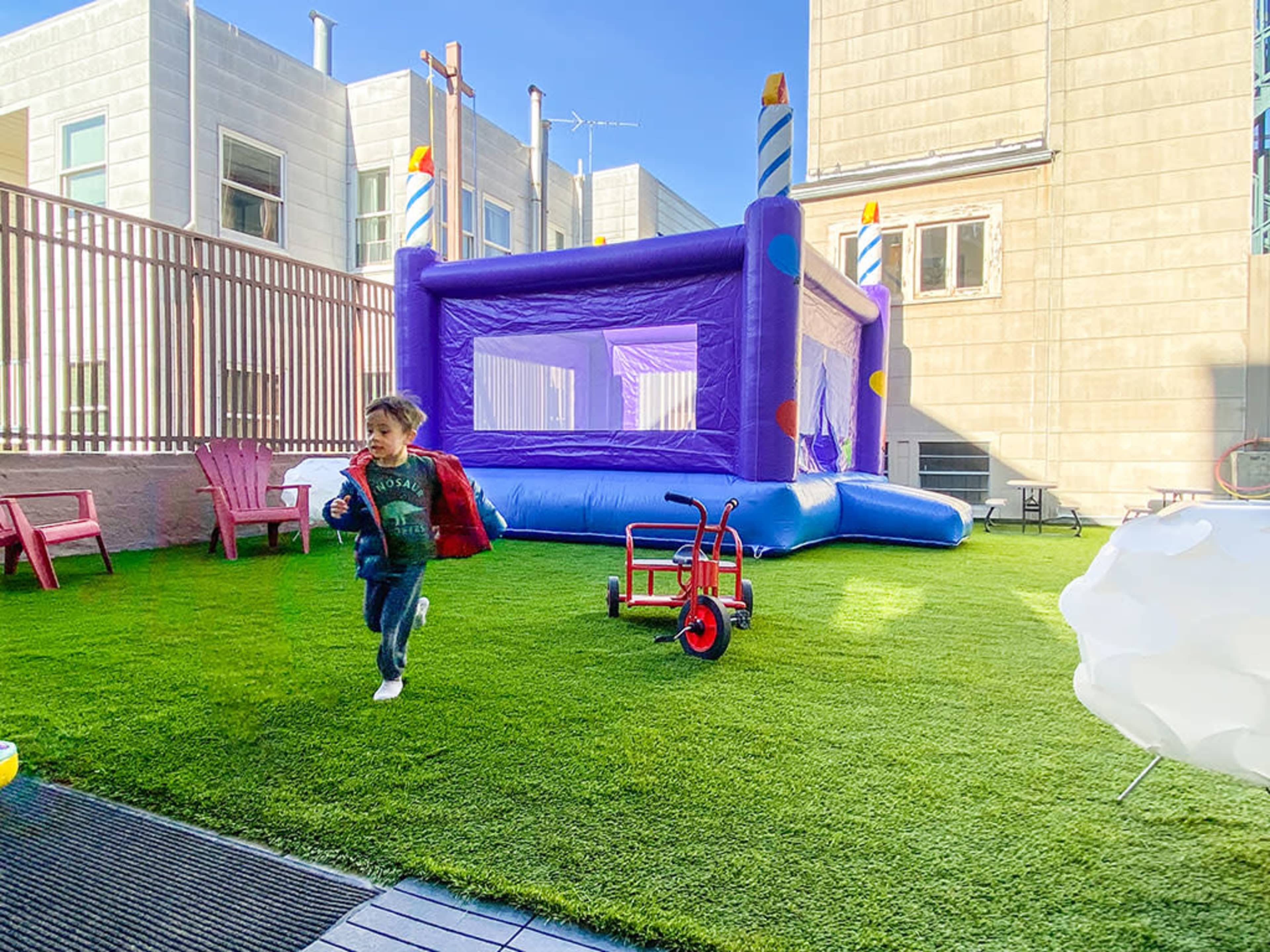 A child runs across a grassy area with a purple bounce house and a red tricycle nearby.