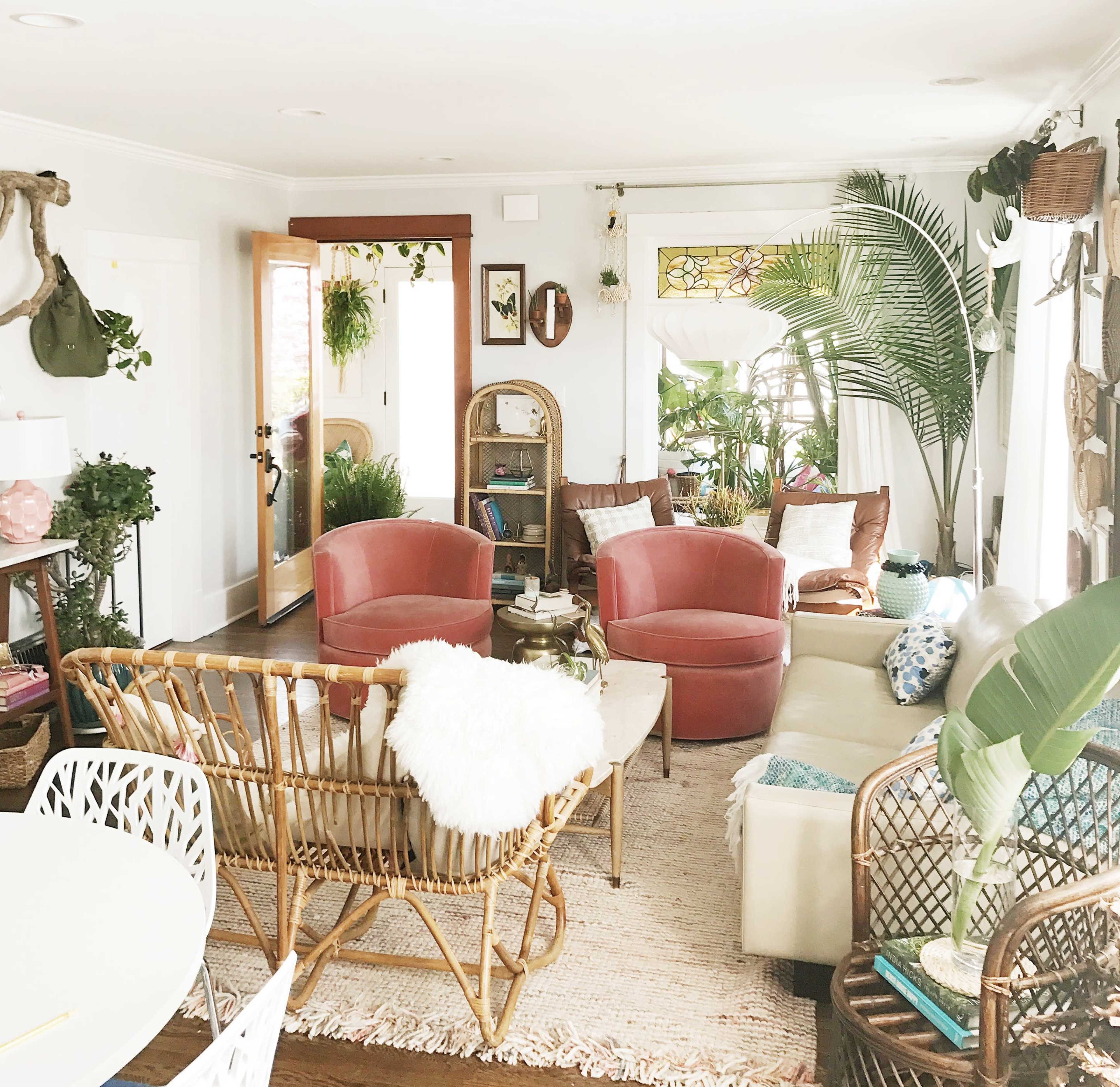 The living room features a mix of modern and vintage furniture, including pink armchairs, a rattan sofa, and a variety of indoor plants.