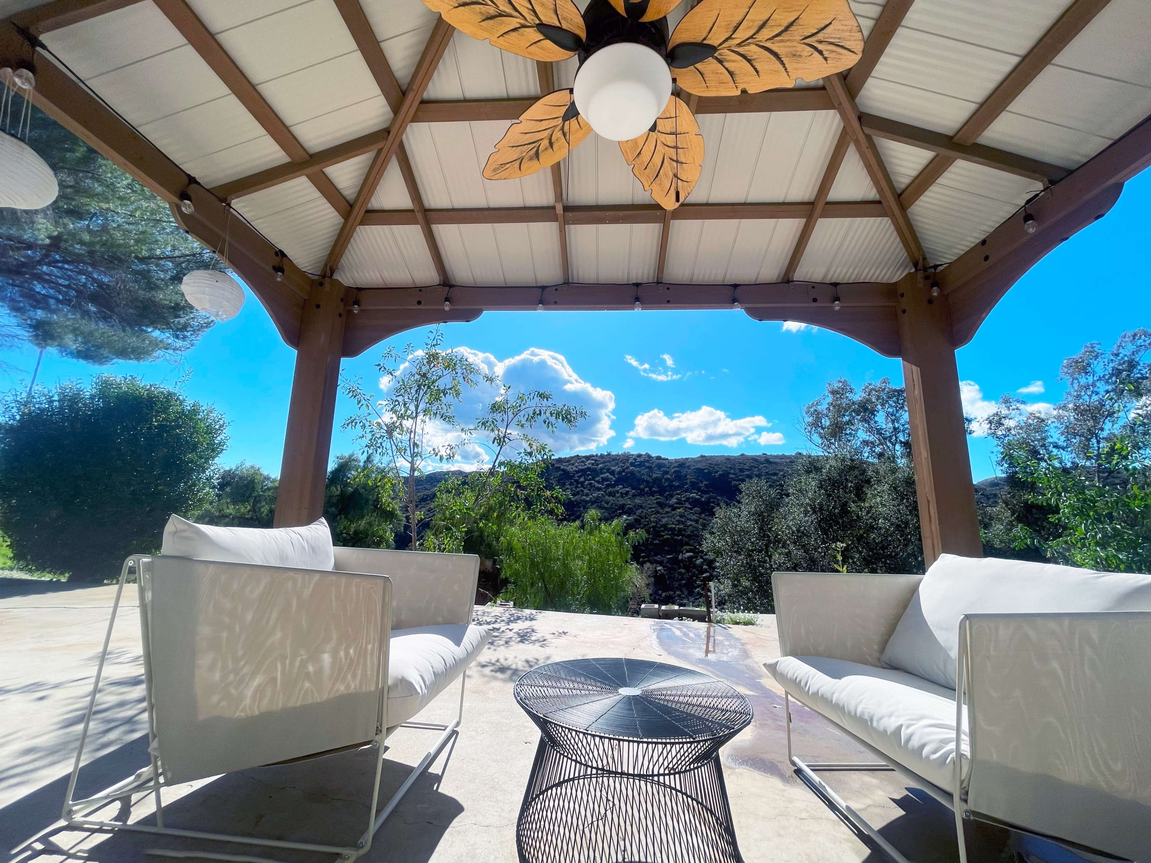 A covered outdoor seating area features two white chairs and a black wire table, overlooking a landscape with trees and hills under a blue sky.