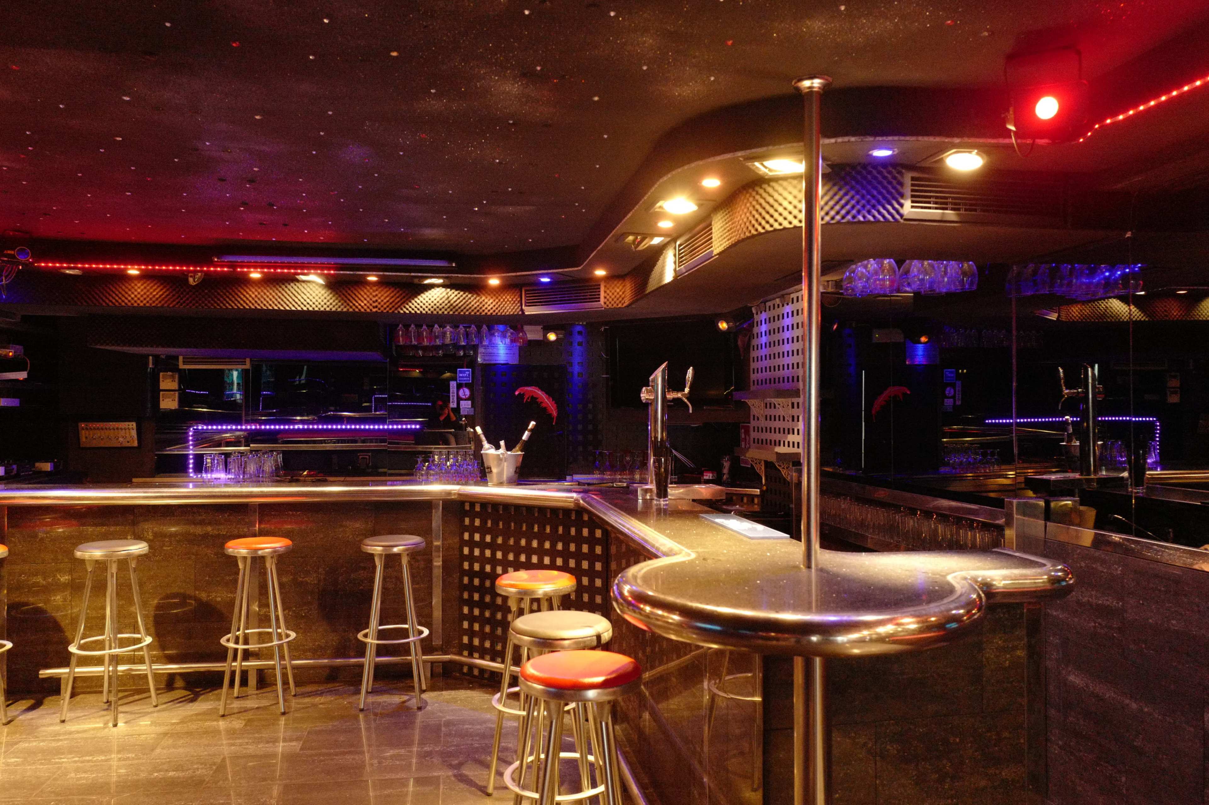 A dimly lit bar with a sleek counter, several metal stools, and colorful LED lighting accentuating the space.
