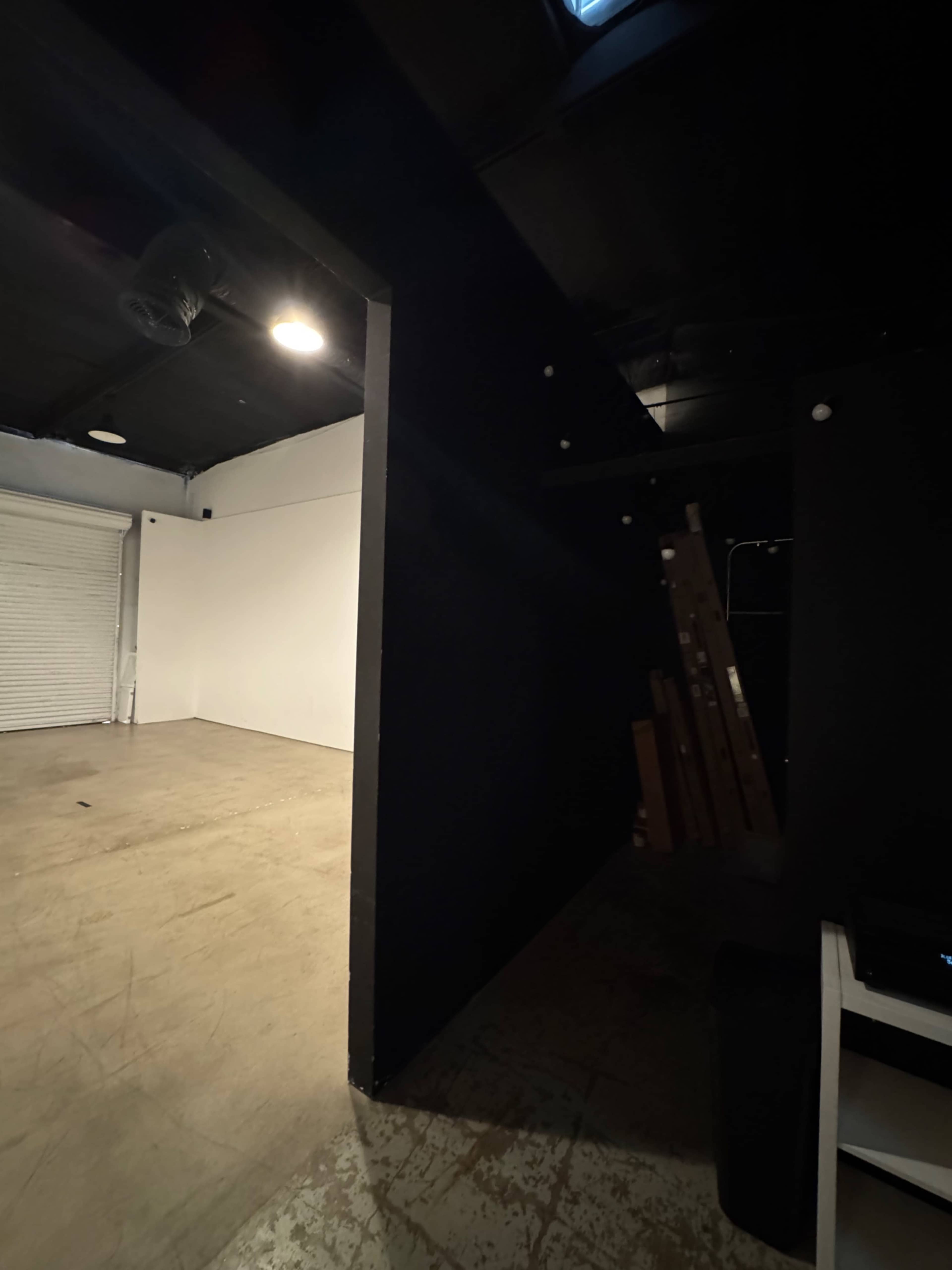 The image shows a spacious, empty room with a partially visible black wall leading to a darkened corridor and a storage area in the corner.