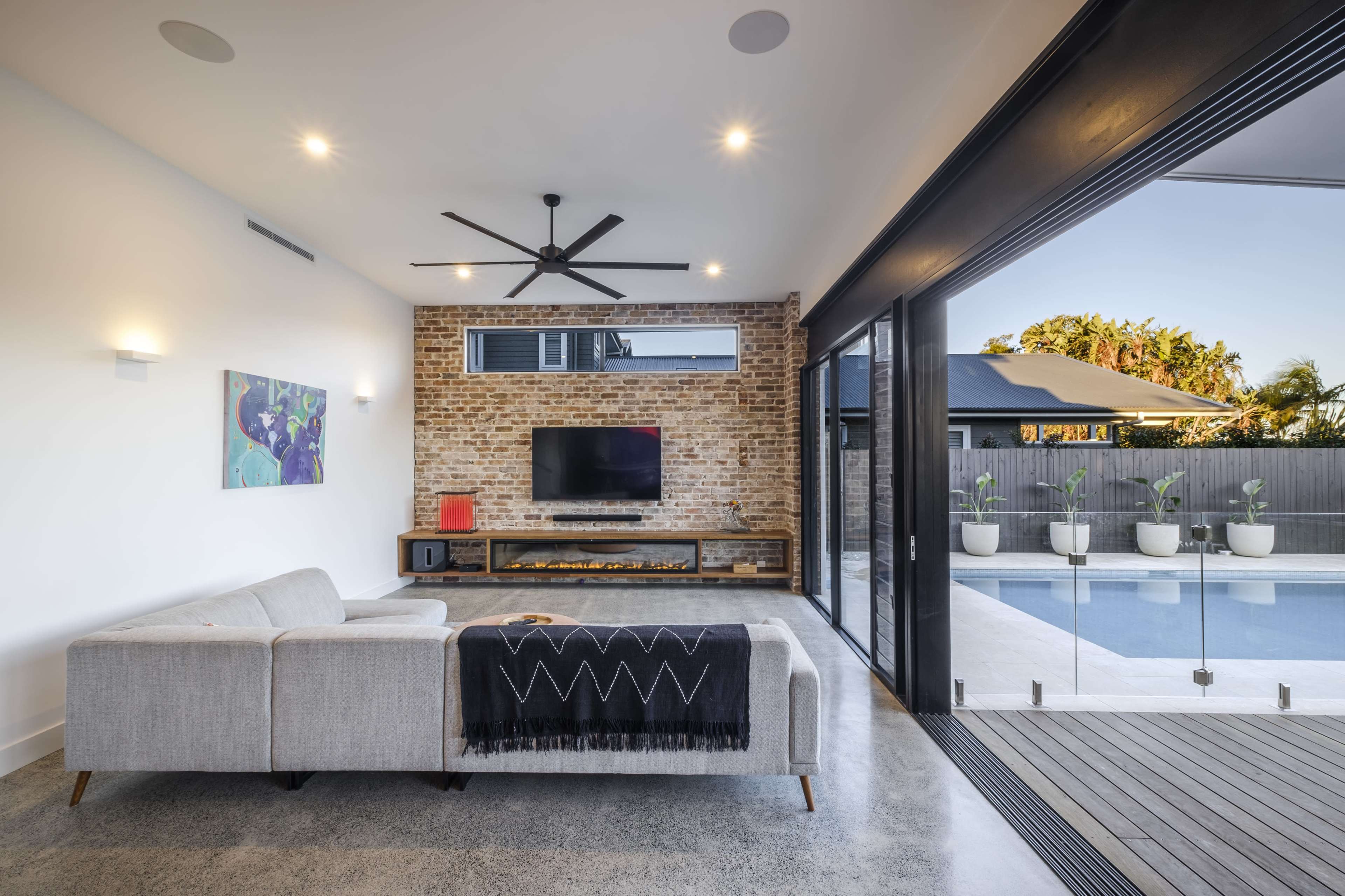 A modern living room features a gray sectional sofa, a brick accent wall with a television, and large sliding glass doors that open to a pool area.