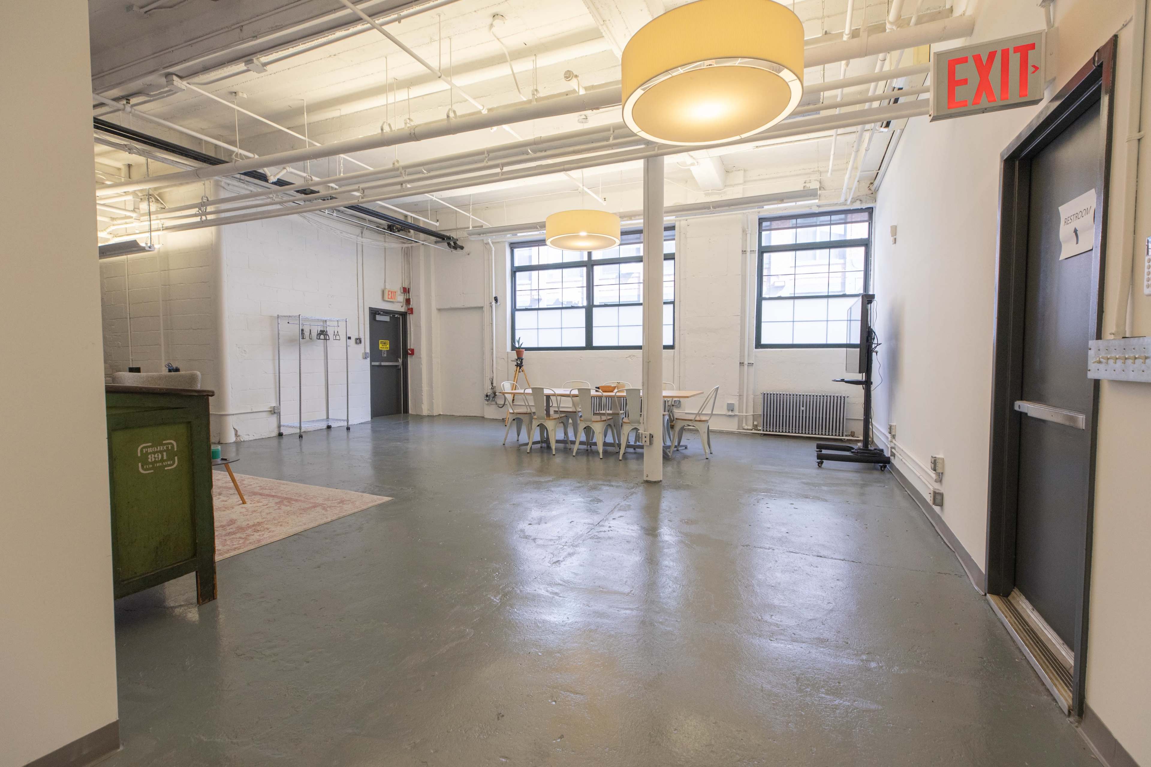 The image shows a spacious, empty room featuring a long table with several chairs, large windows, and an exit sign.