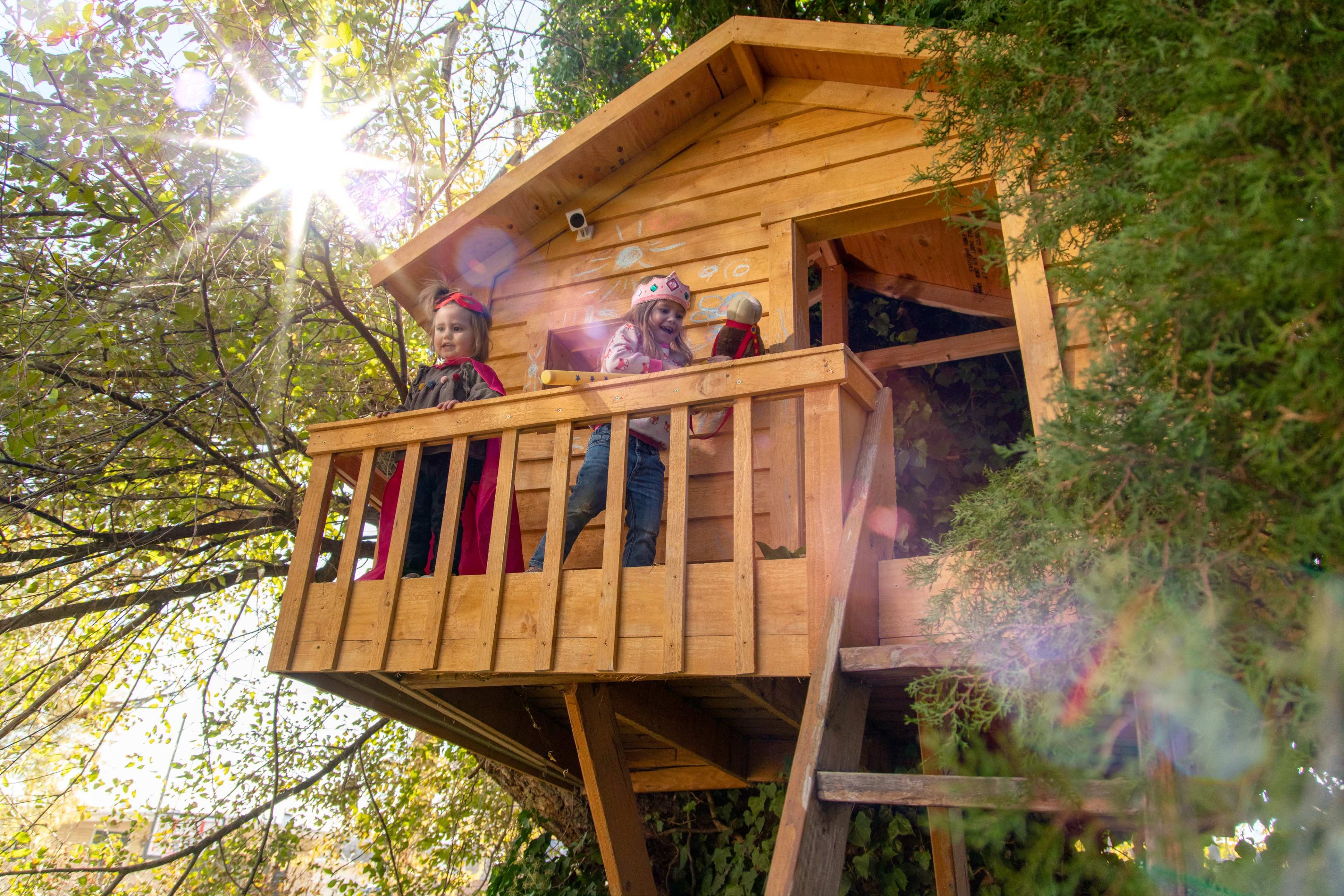 Two children stand on the balcony of a wooden treehouse surrounded by greenery and sunlight.