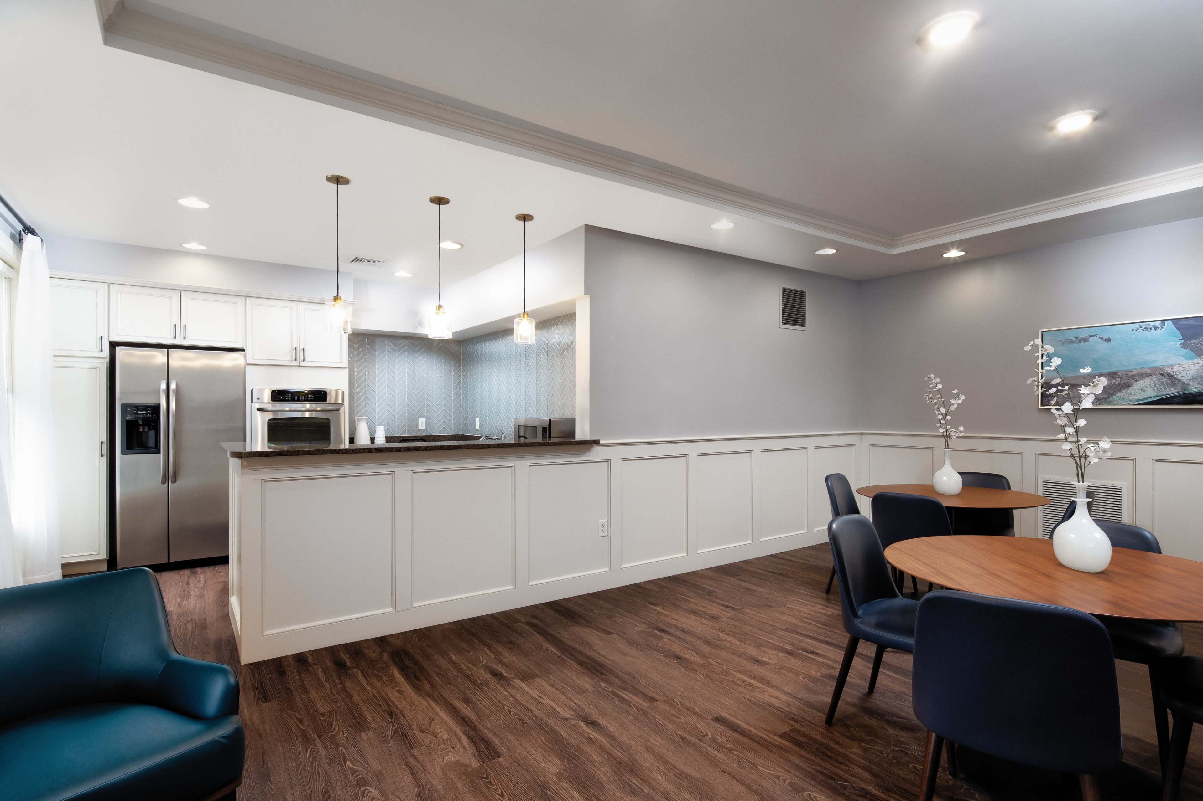 A modern kitchen and dining area with a light gray wall, wooden flooring, stainless steel appliances, and round tables with chairs.