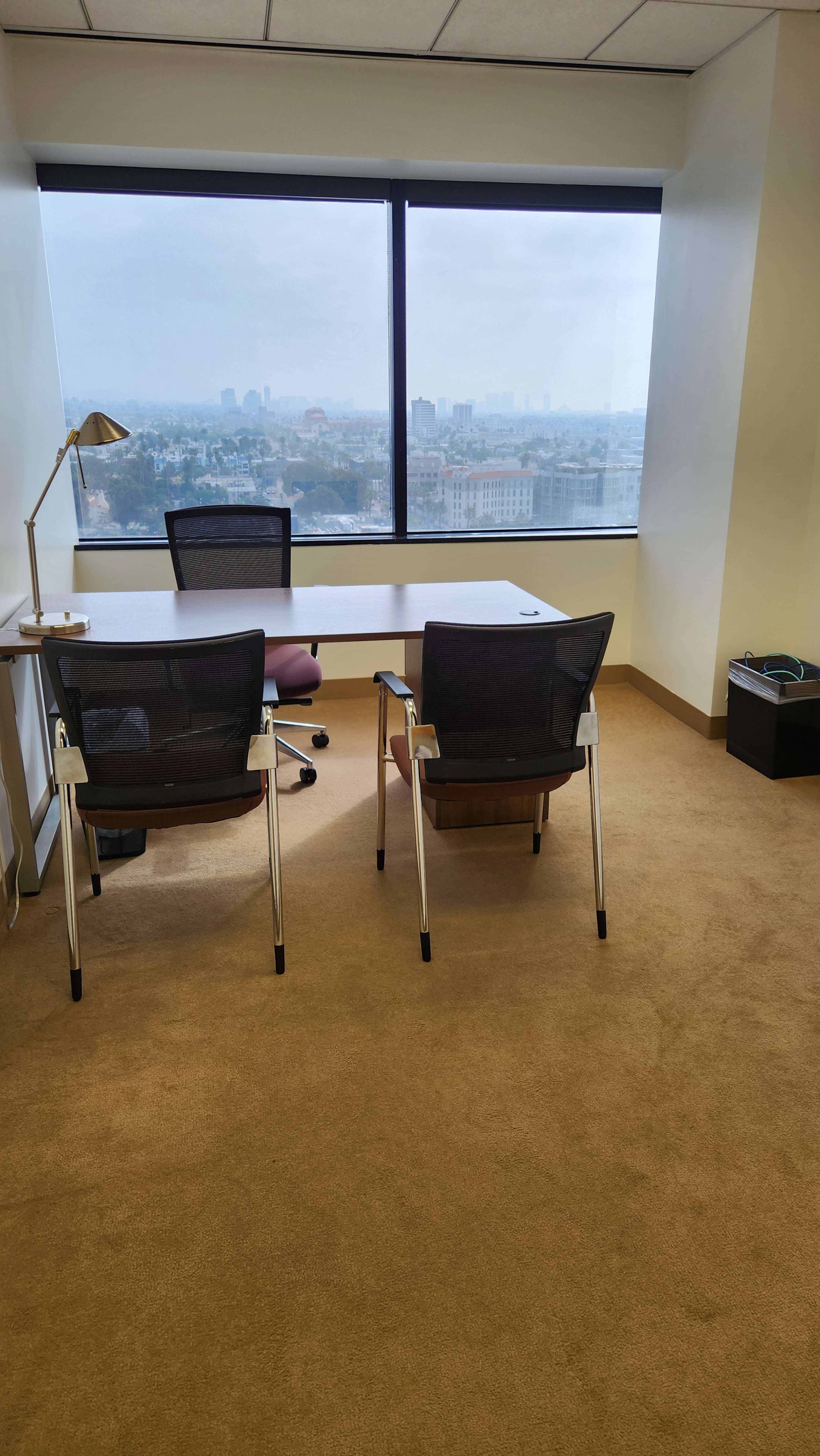 An office space with a large window offering a view of a city skyline, two chairs positioned at a table, and a lamp on the desk.