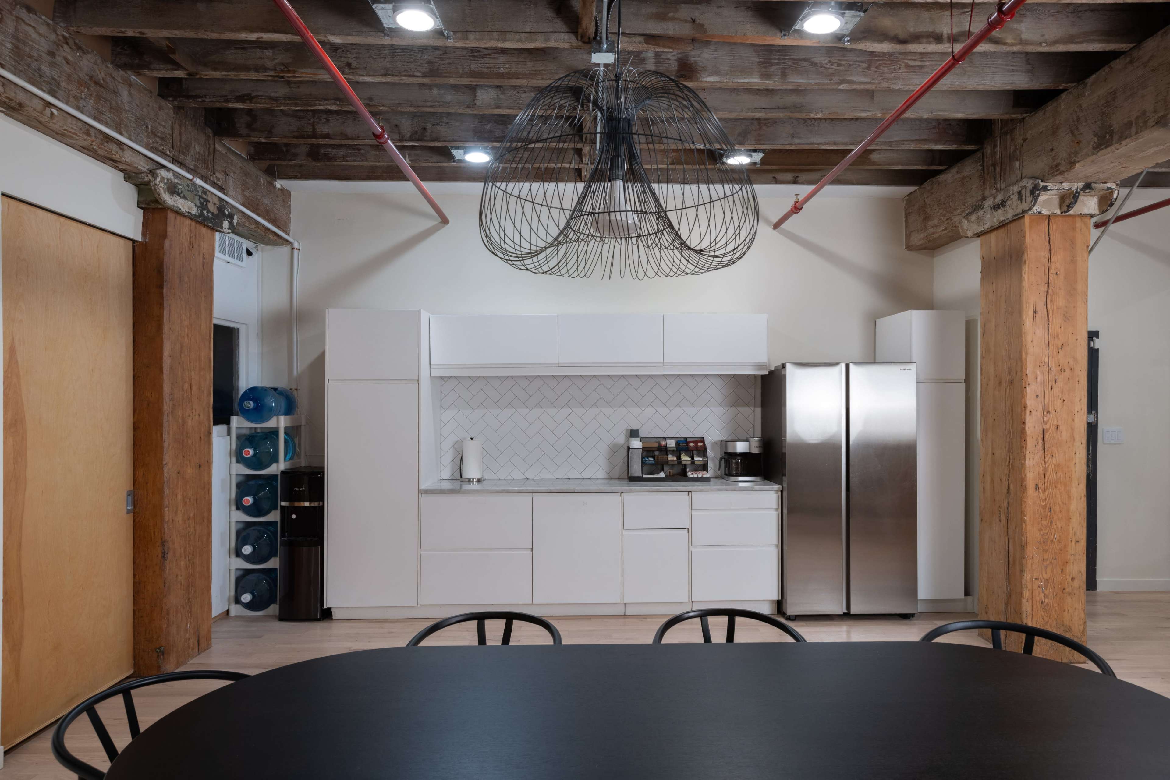 A modern kitchen area features white cabinetry, a stainless steel refrigerator, and a large black dining table under a wire chandelier.