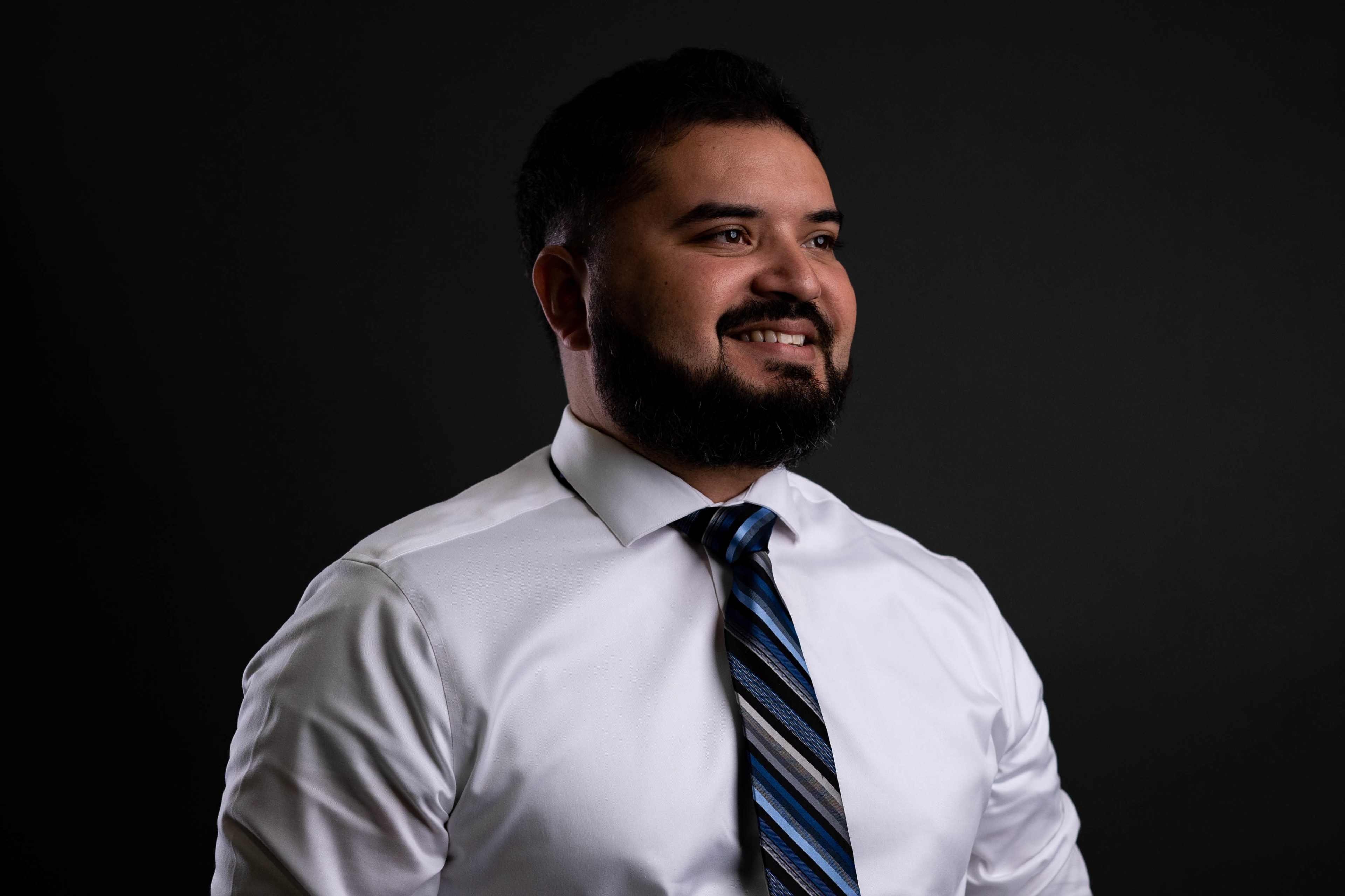 A man with a beard smiles while wearing a white dress shirt and a striped tie against a dark background.
