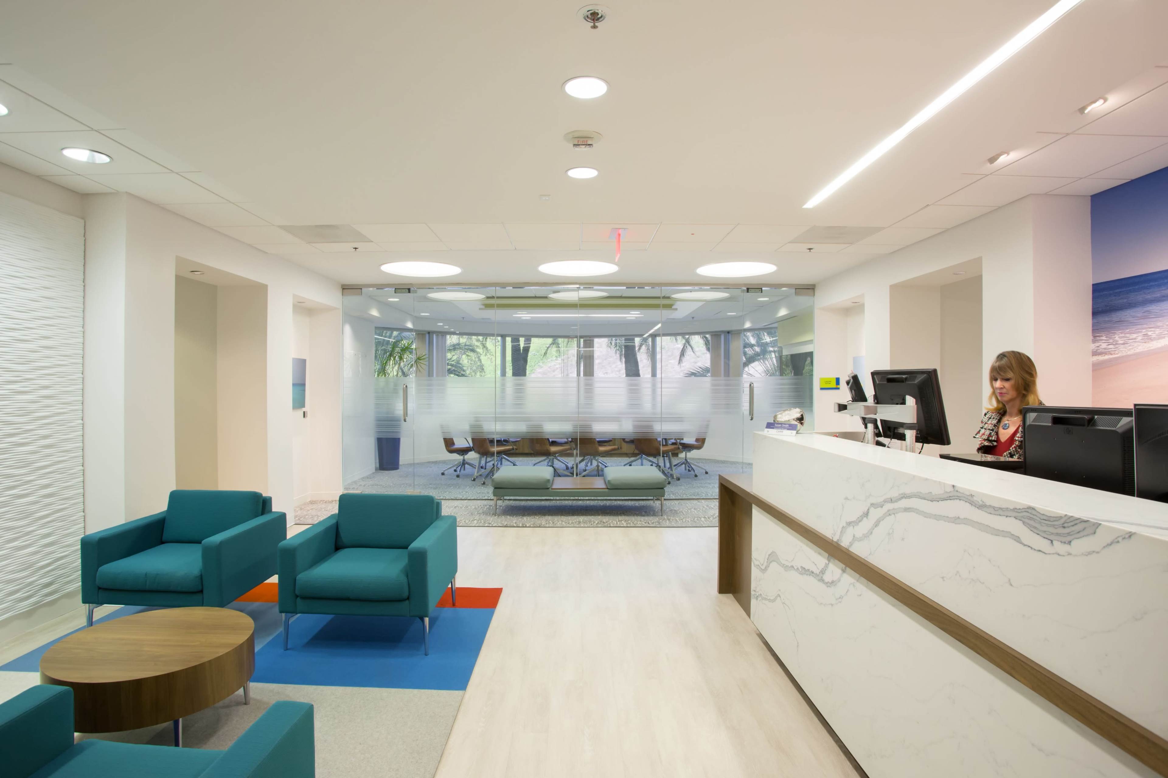 The image shows a modern reception area with teal seating, a stone front desk, and a glass-walled meeting space in the background.
