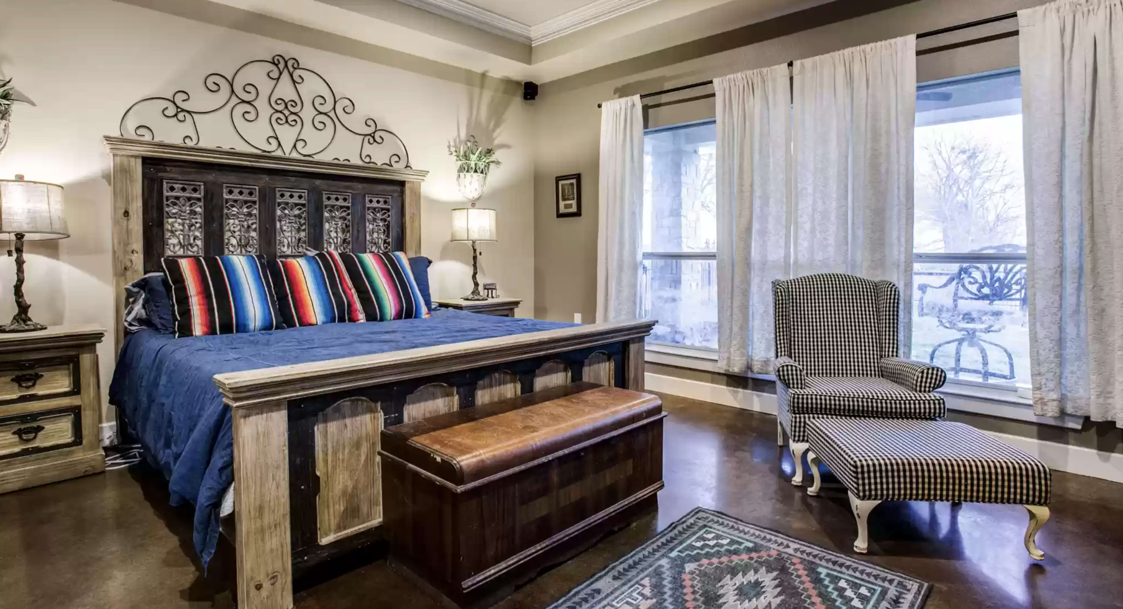 A cozy bedroom with a wooden bed frame, multicolored striped pillows, a decorative headboard, a bench at the foot of the bed, and a patterned armchair beside a window with sheer curtains.