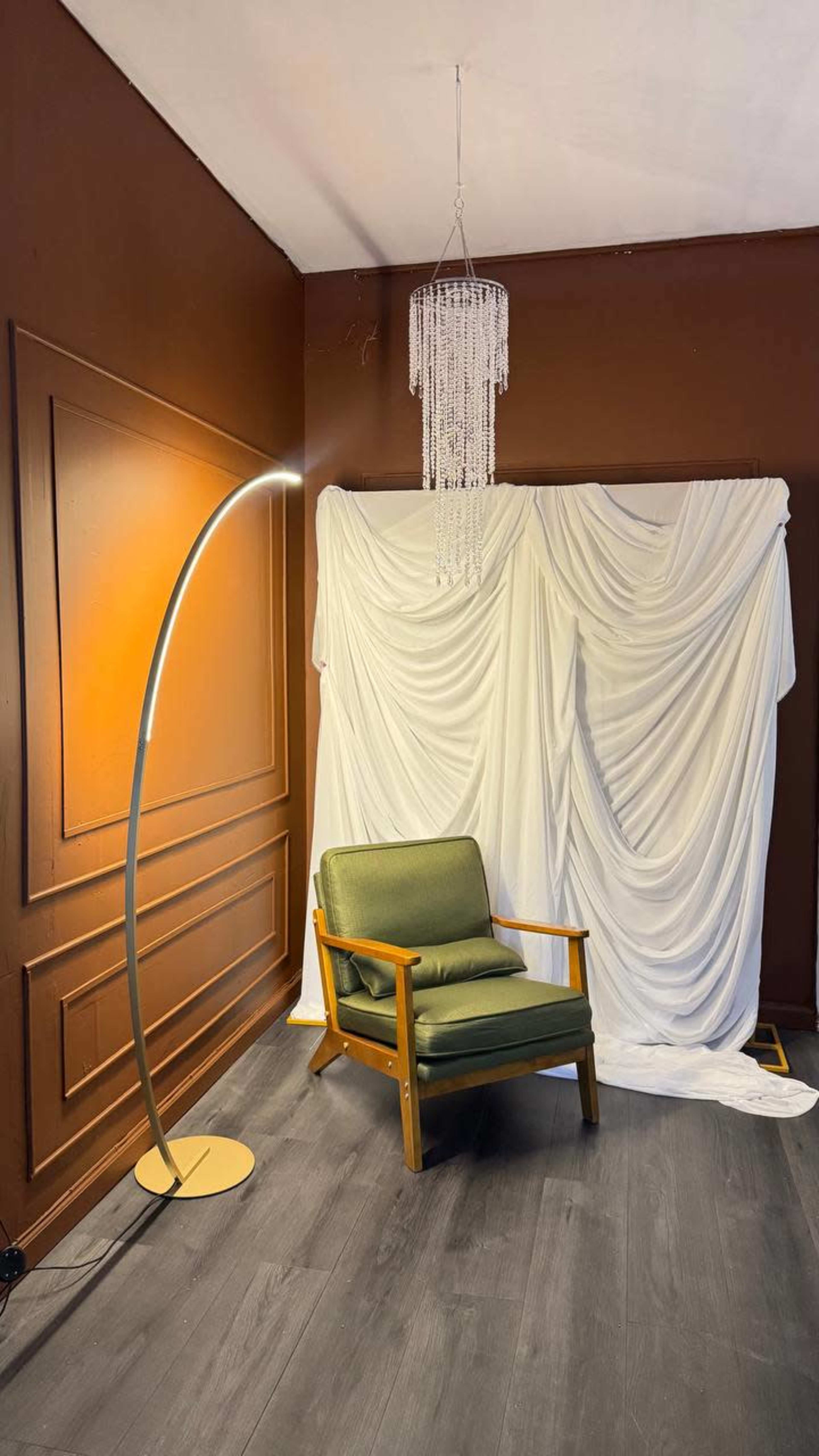 The image shows a green armchair beside a modern arc lamp, with draped white fabric and a chandelier in the background.
