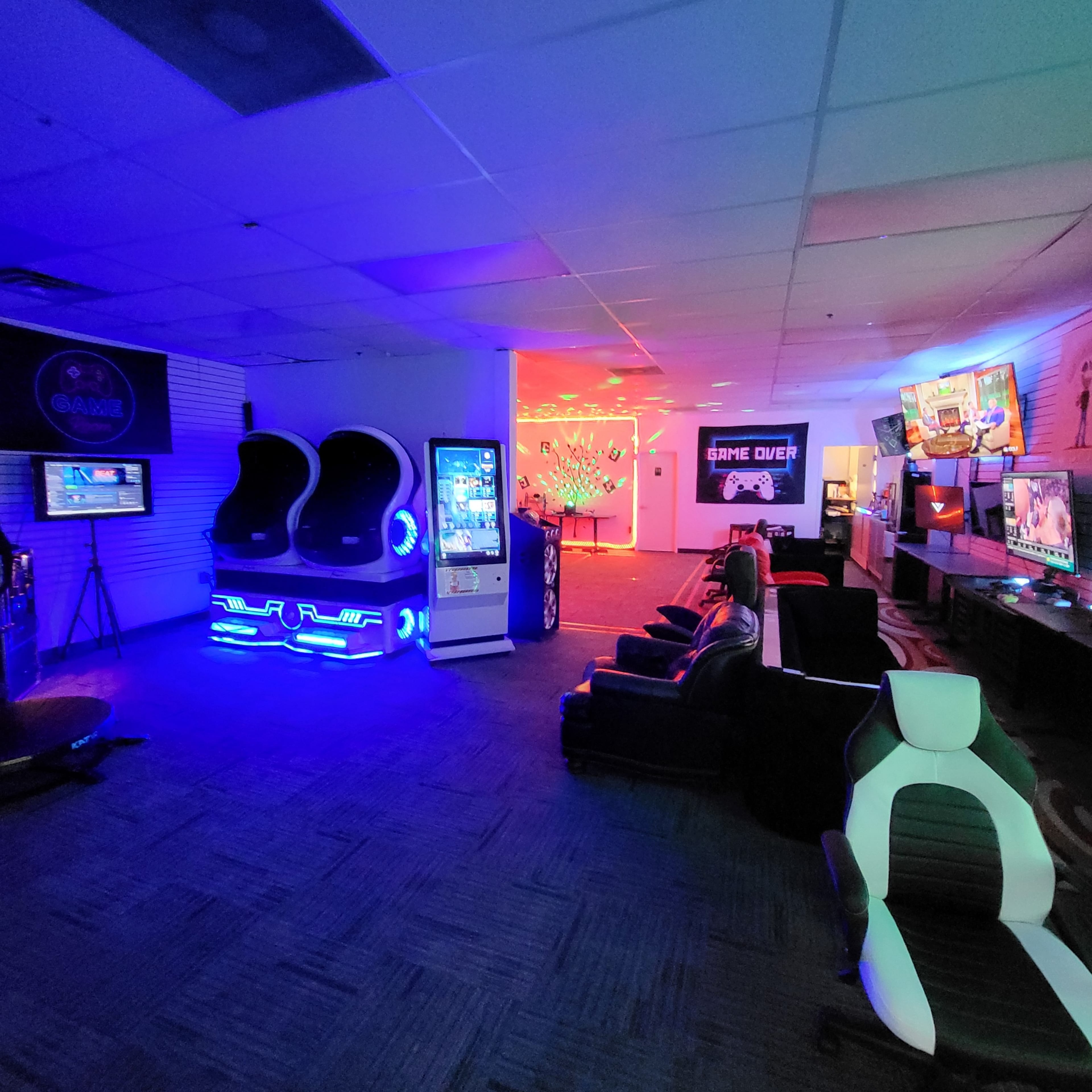 The image shows a gaming lounge featuring arcade machines, multiple gaming consoles, and various seating arrangements illuminated by colorful LED lighting.