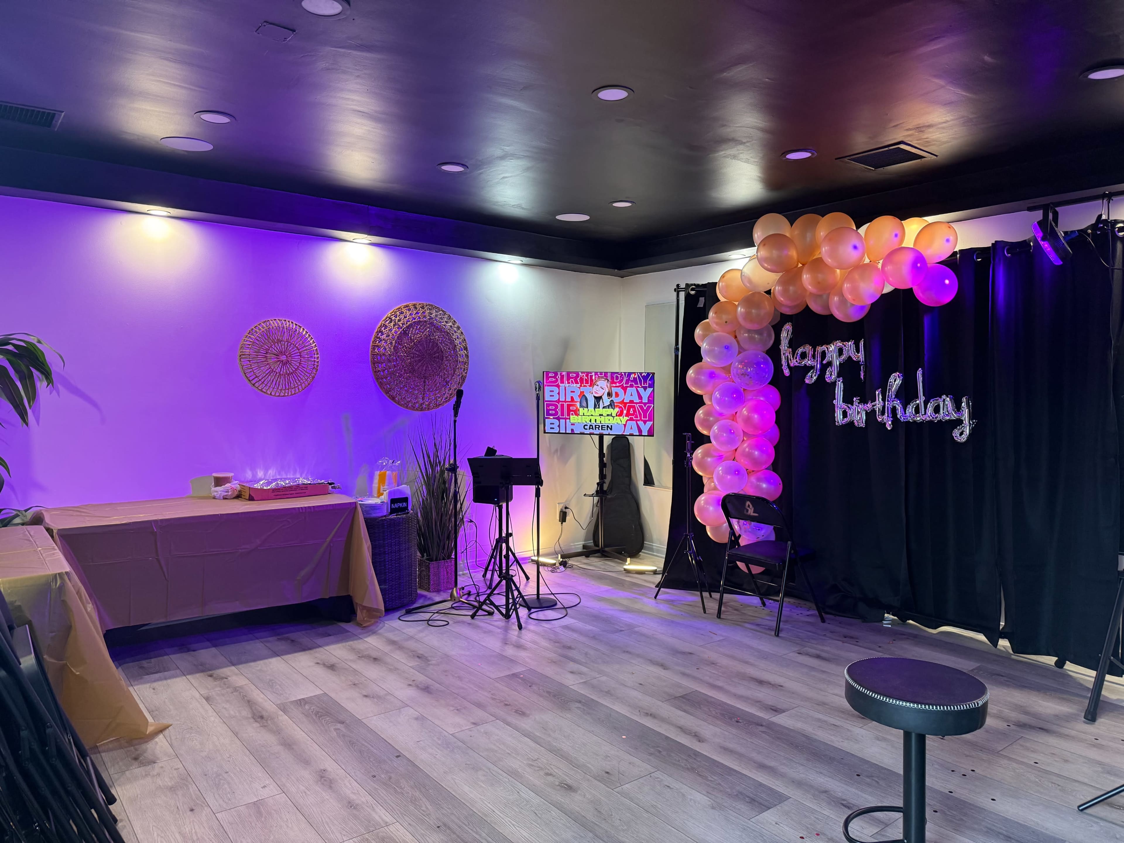 The image shows a decorated event space for a birthday celebration, featuring balloons, a lighted sign, a table with party supplies, and a black curtain backdrop.