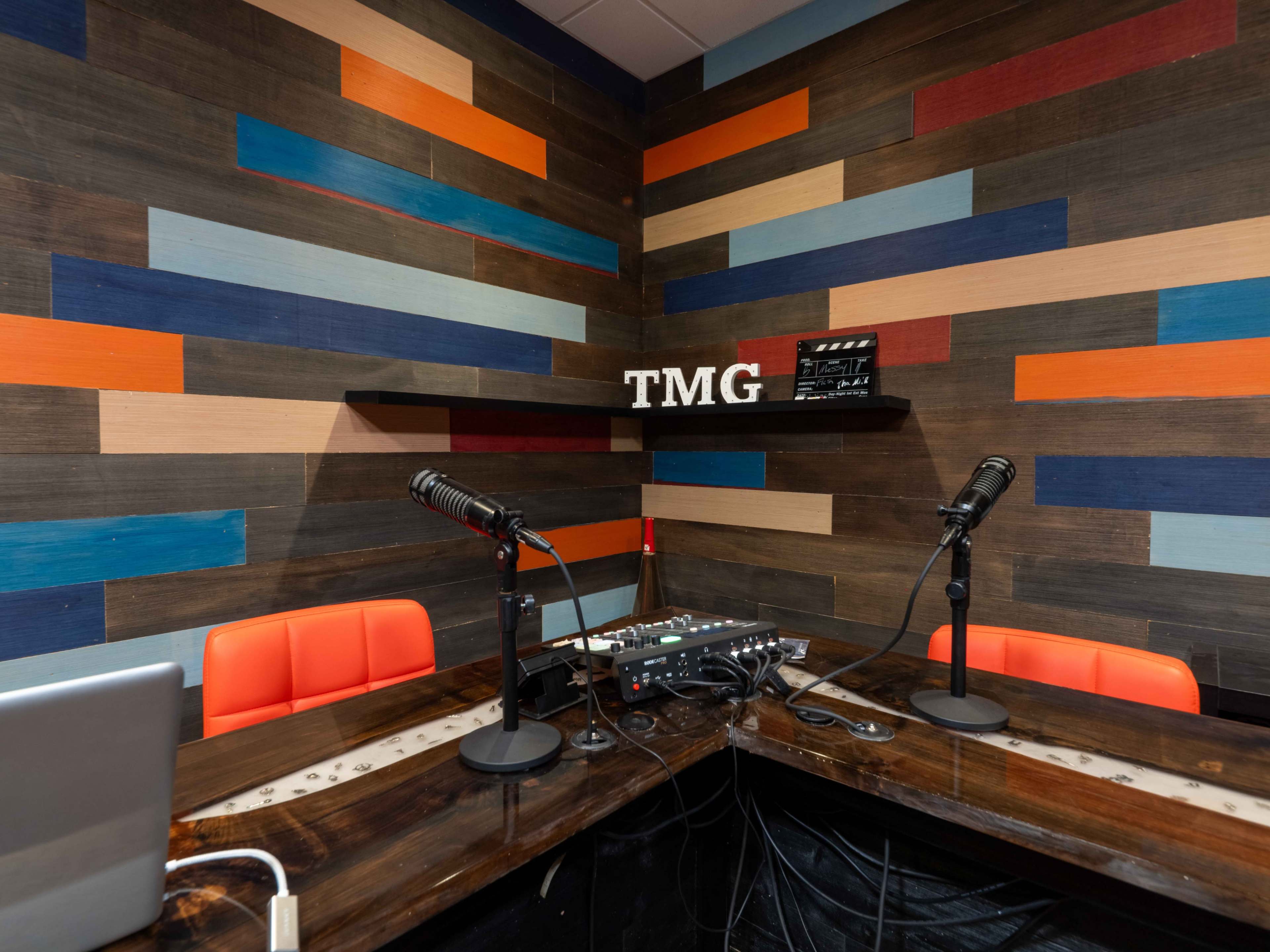 A recording studio with a wooden wall featuring colorful stripes, two microphones on a table, and a sign that reads "TMG."