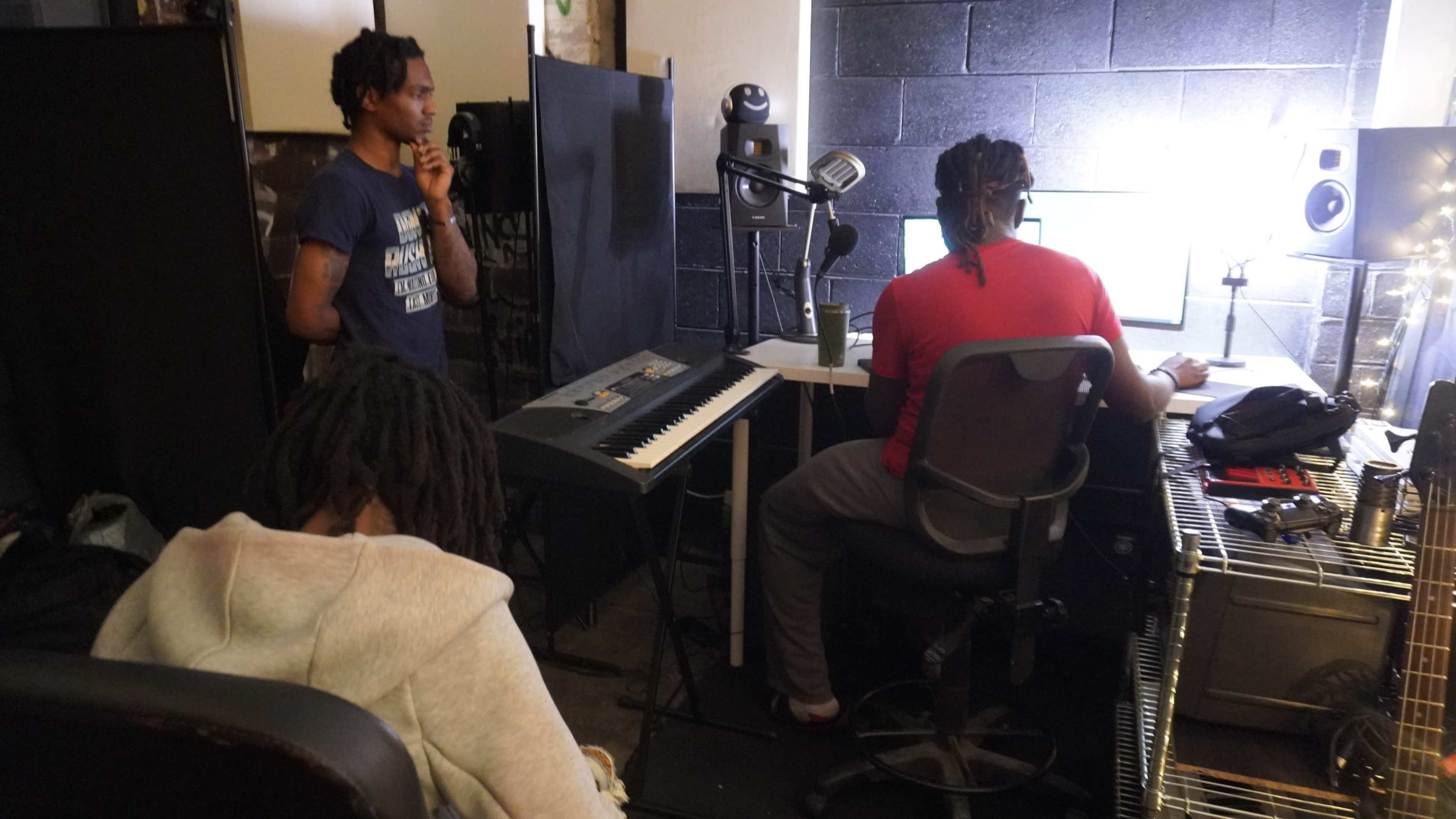 Three people are in a recording studio, with one seated at a keyboard and computer, another seated with their back to the camera, and a third standing and observing.