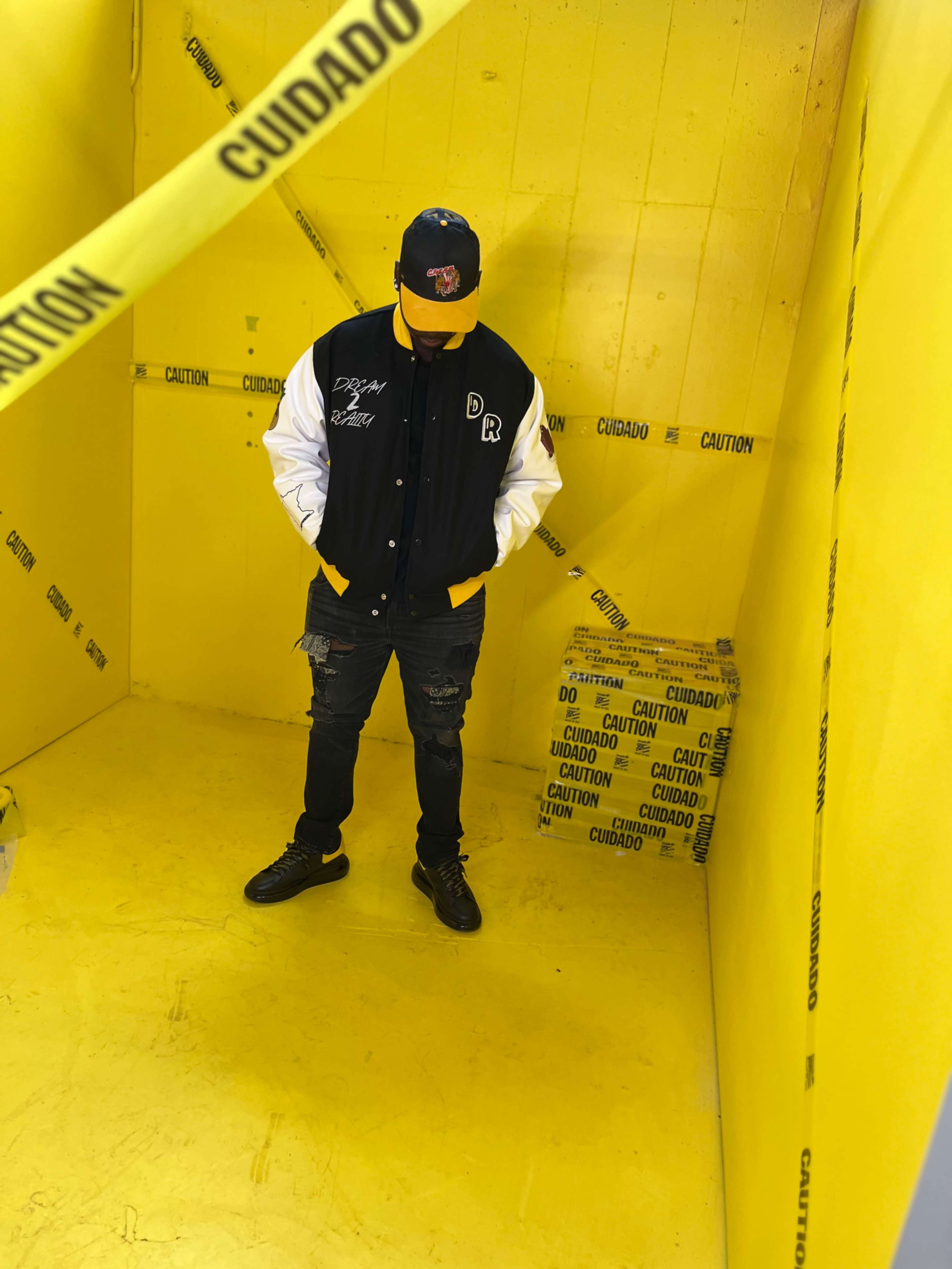 A person in a black and white jacket and cap stands in a yellow room with caution tape and a cardboard box.
