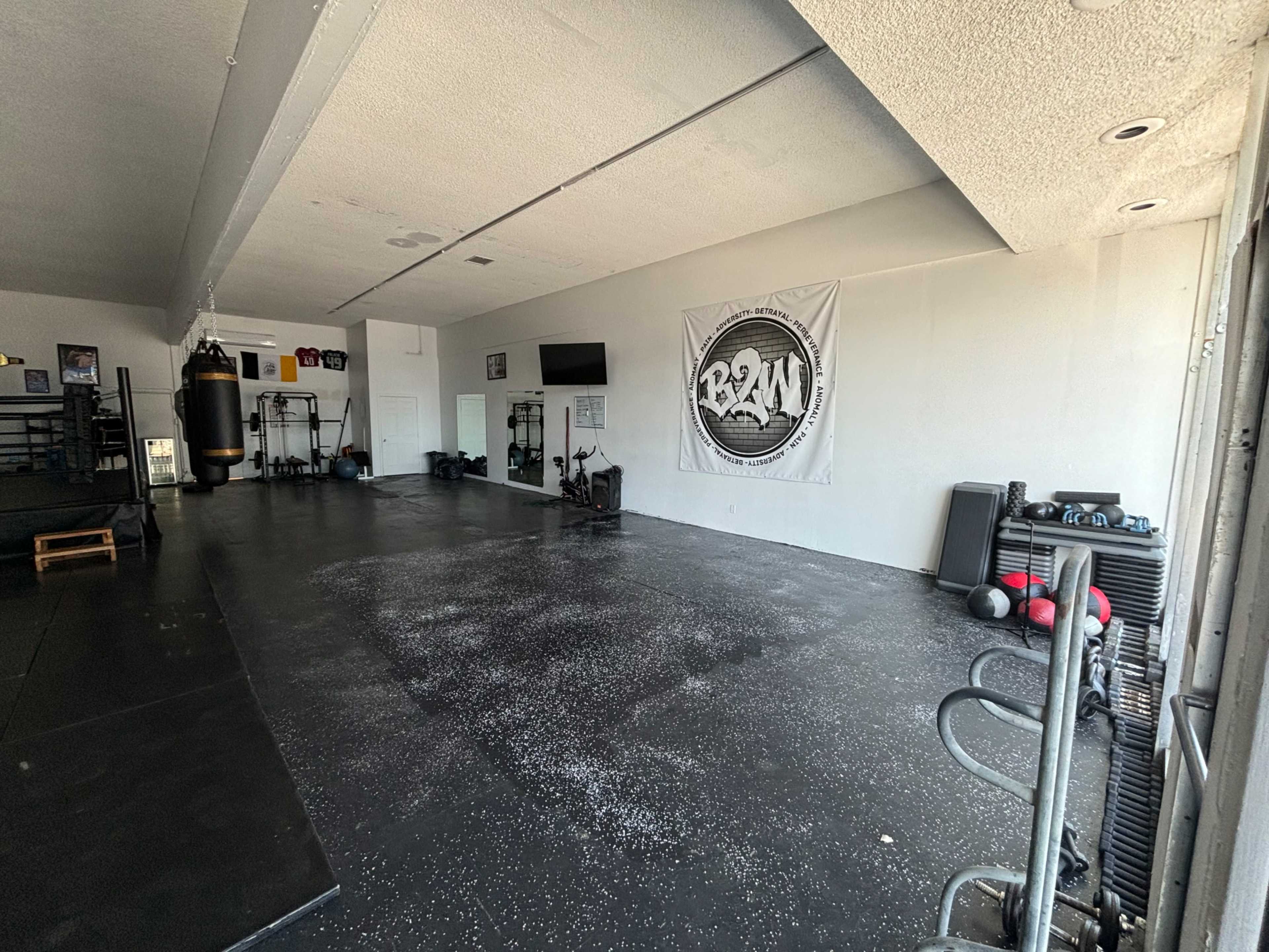 The image shows an empty gym space with black flooring, a punching bag, various fitness equipment, and a large wall-mounted banner.