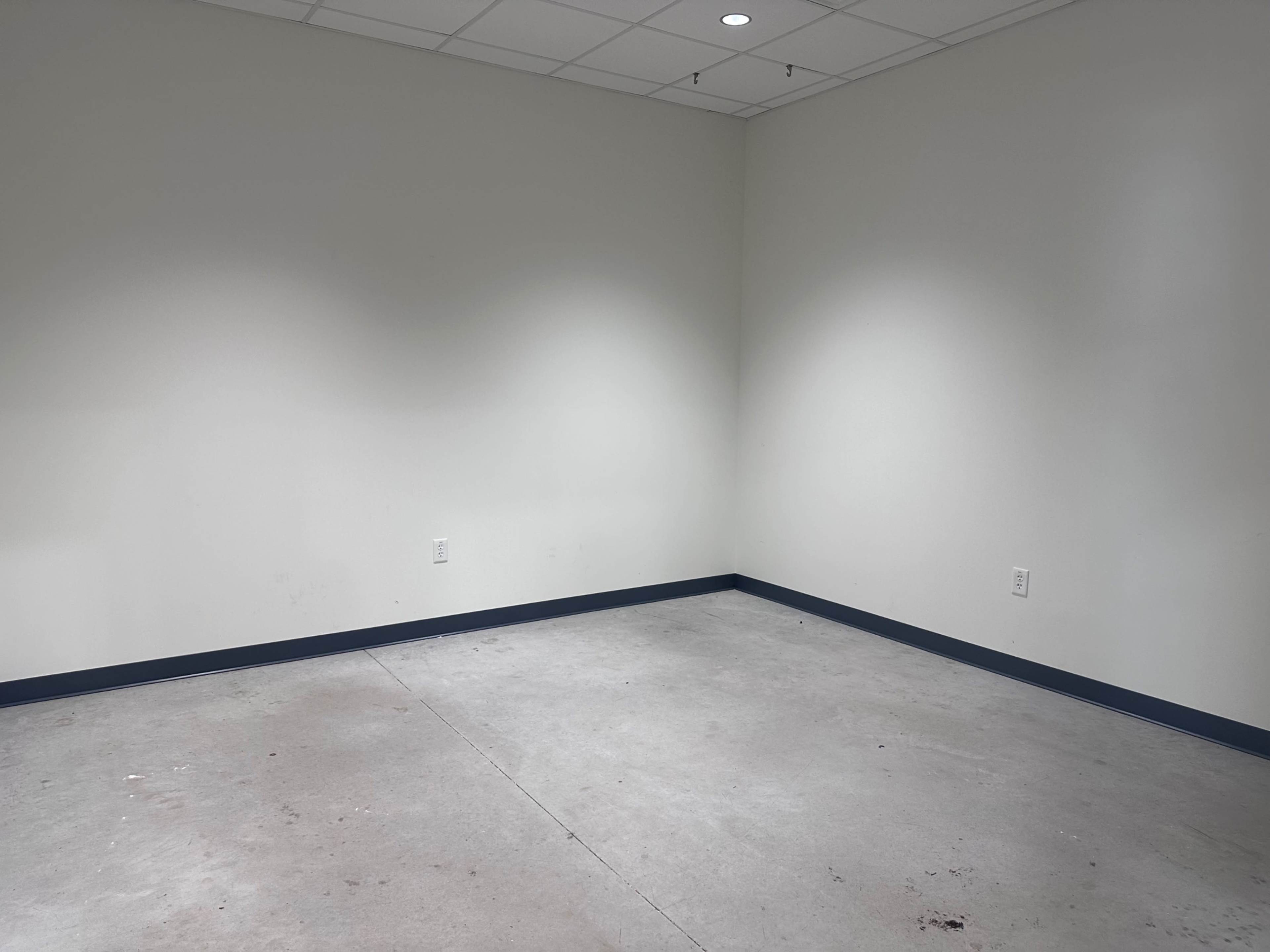 The image shows a vacant room with plain white walls and a gray concrete floor.