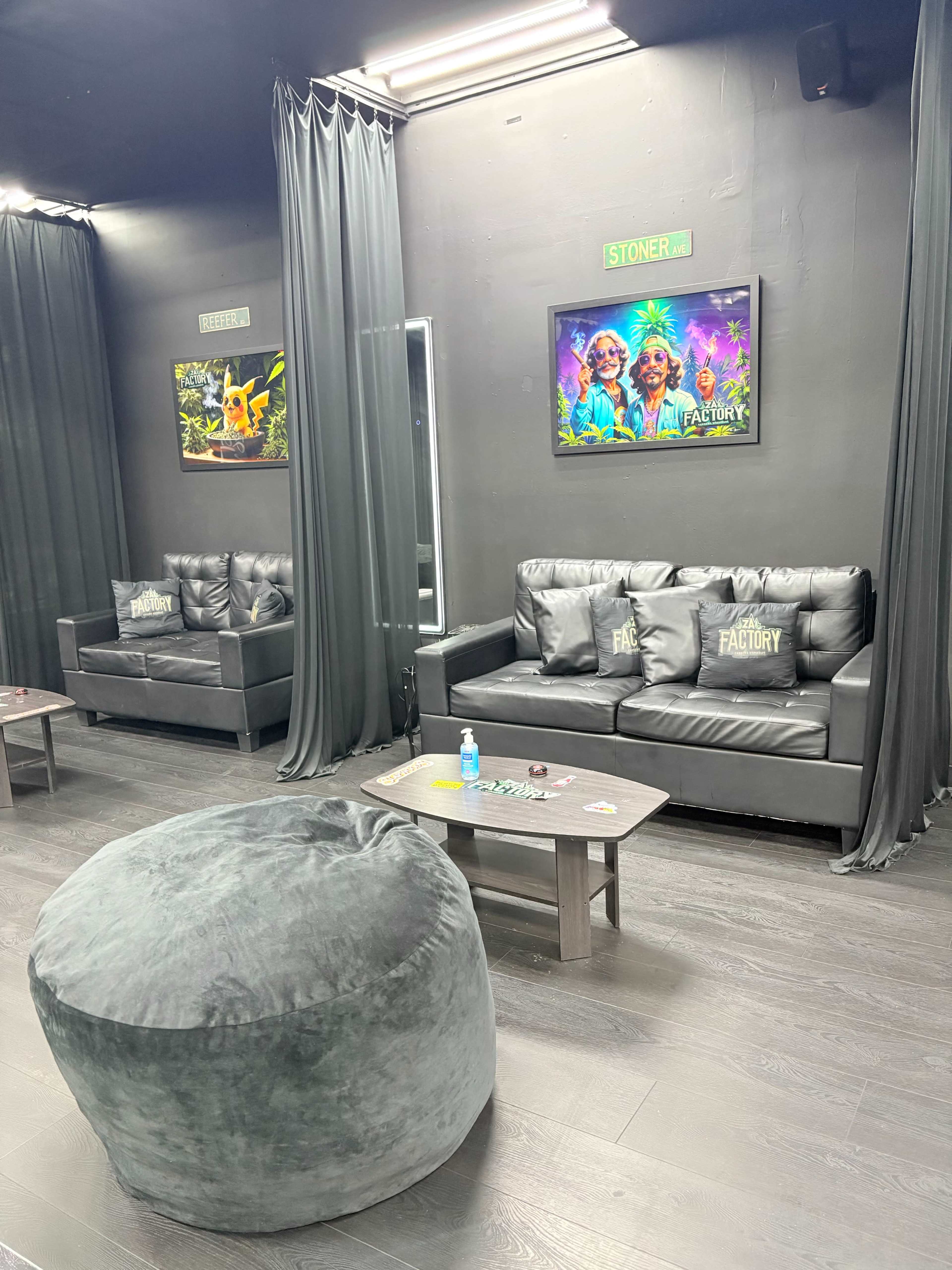 The image shows a lounge area featuring black sofas, a gray bean bag, and framed colorful artwork on the walls, with curtains separating the spaces.