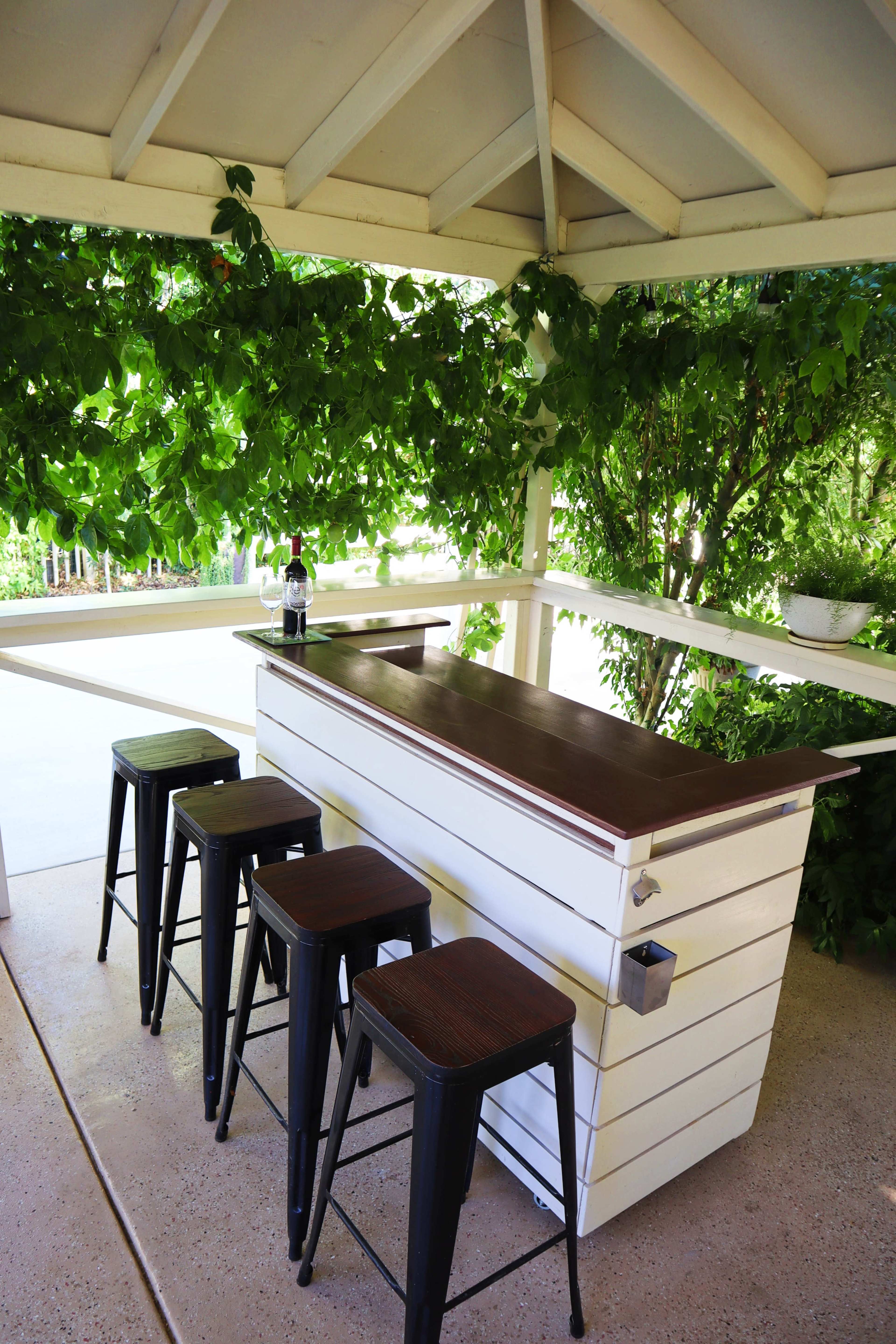 A outdoor bar with a wooden countertop is surrounded by greenery and accompanied by four black metal stools.