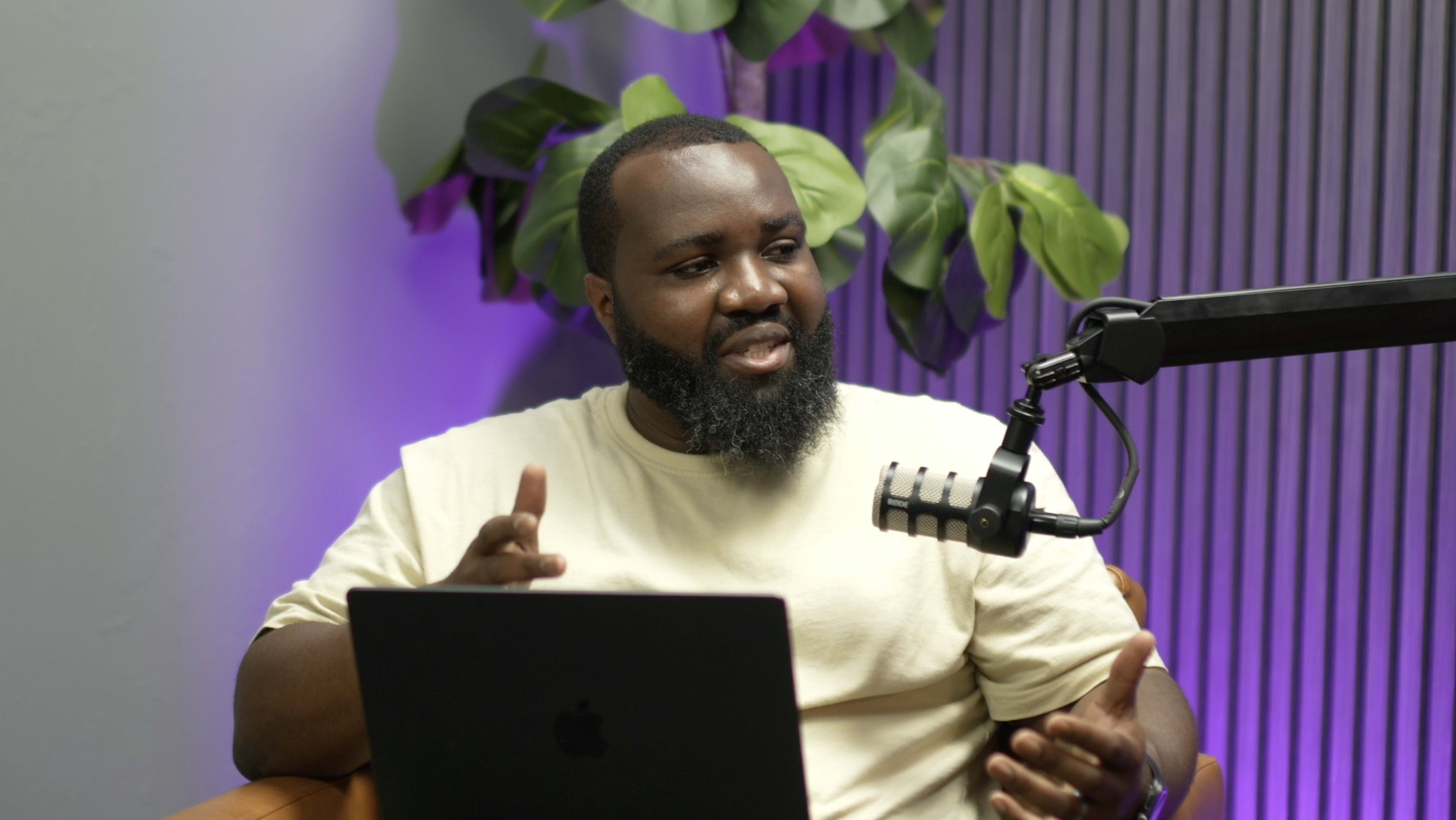 A man with a beard gestures while speaking into a microphone, seated in front of a laptop against a backdrop of plants.