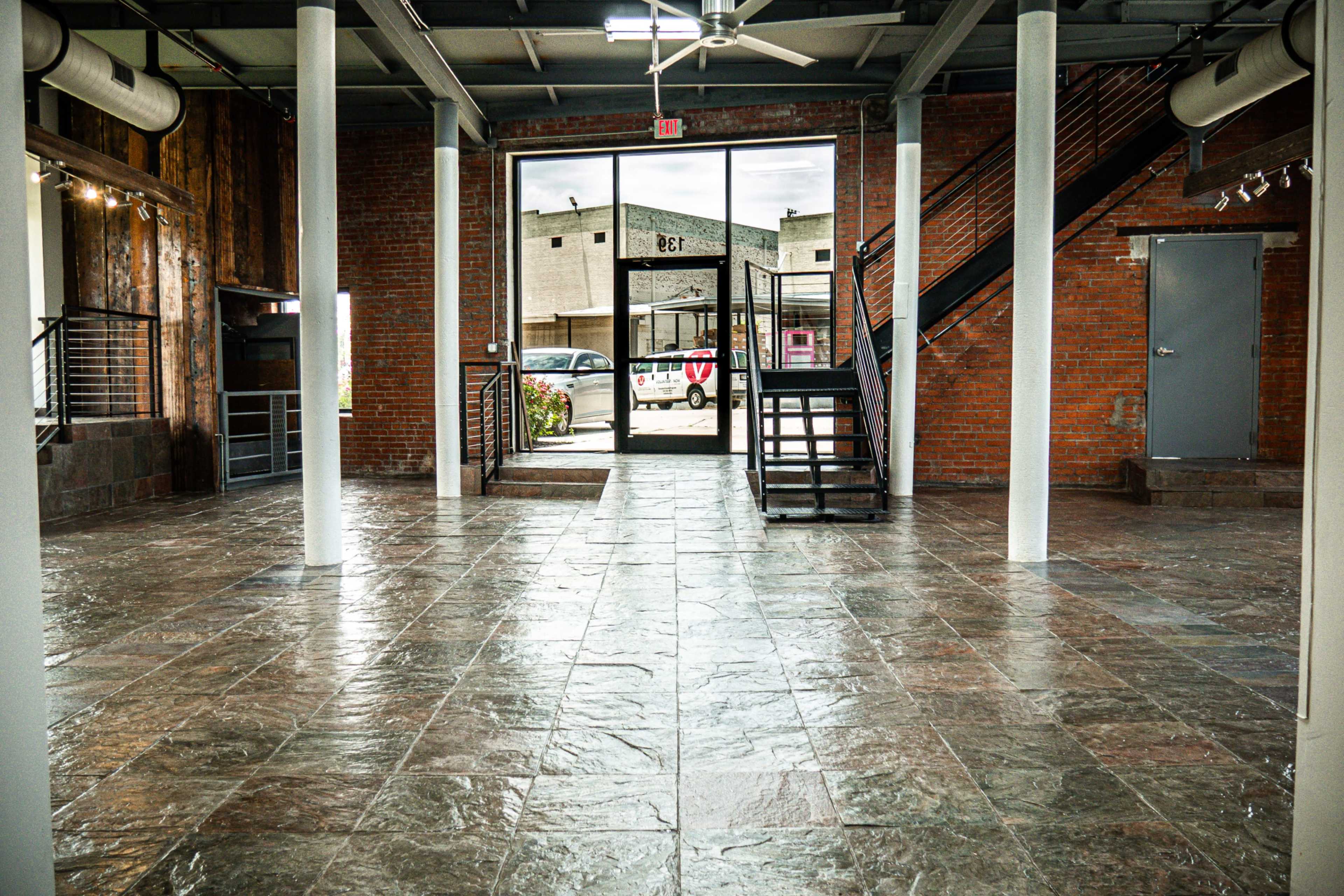 The image shows an empty industrial-style interior space with a tiled floor and large windows leading to an outdoor area.
