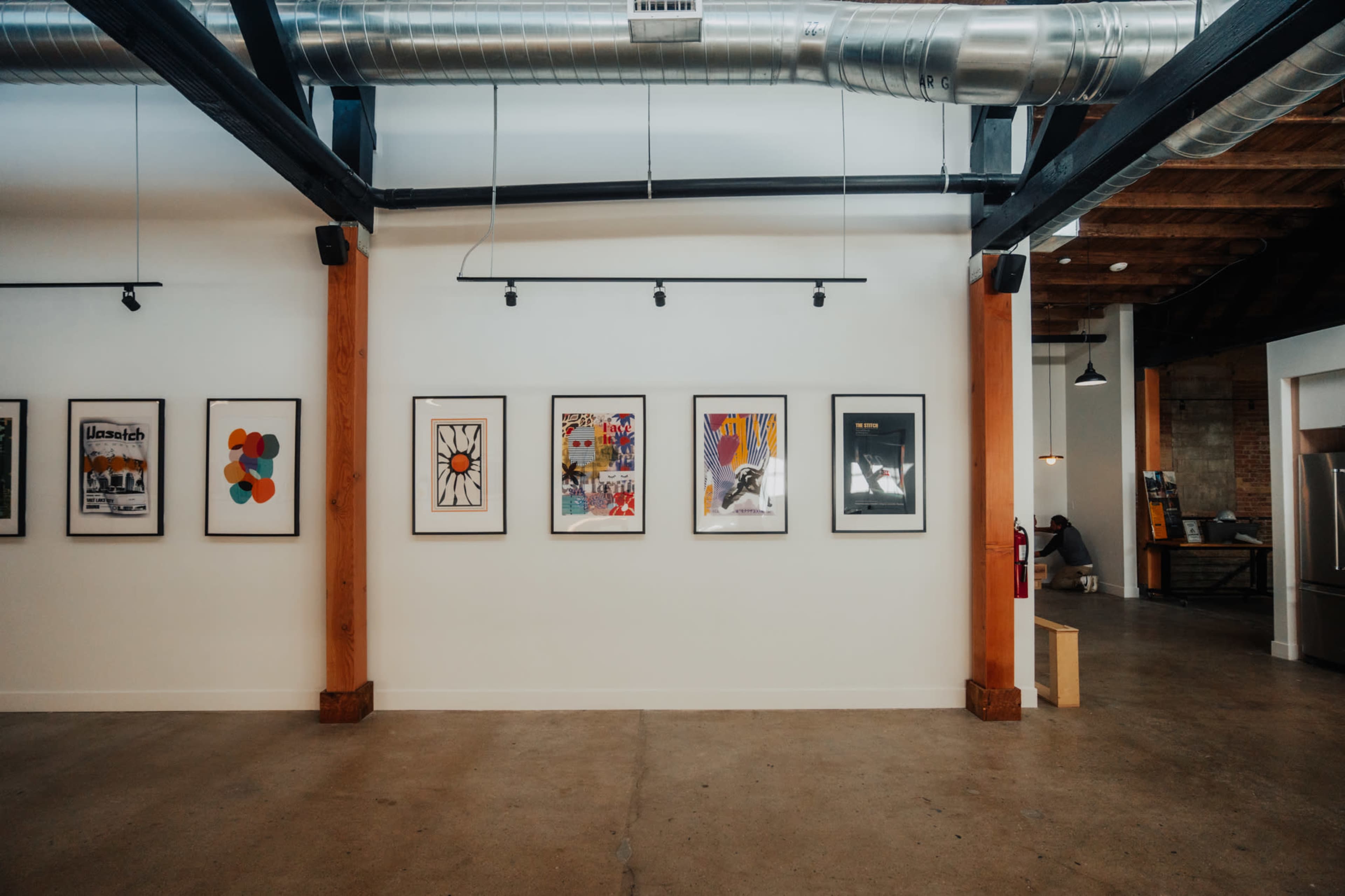 The image shows a gallery wall featuring a series of framed artworks in an industrial-style room with exposed beams and ducts.