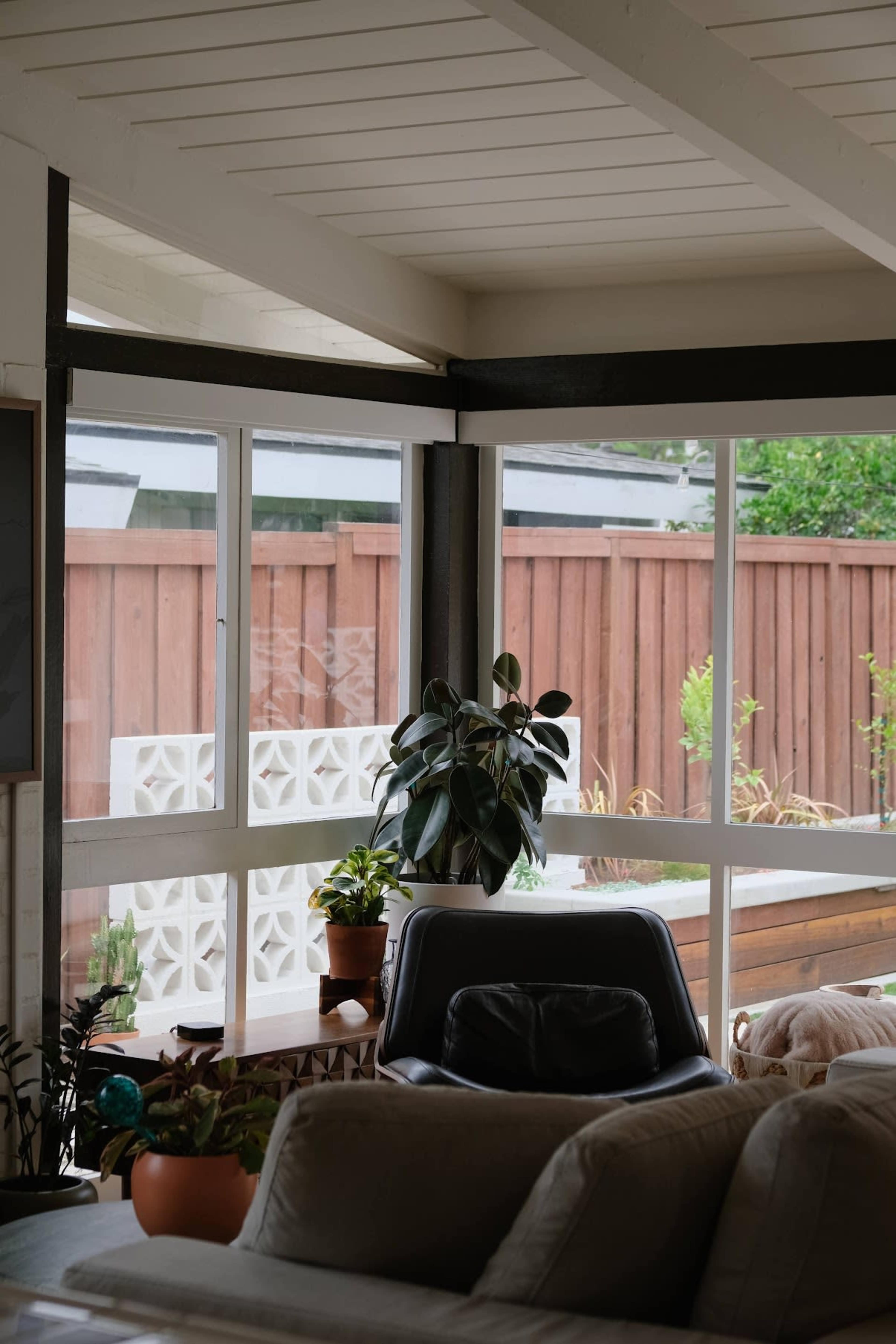 The image depicts a cozy living room with a large window facing a wooden fence, featuring plants both inside and outside the window.
