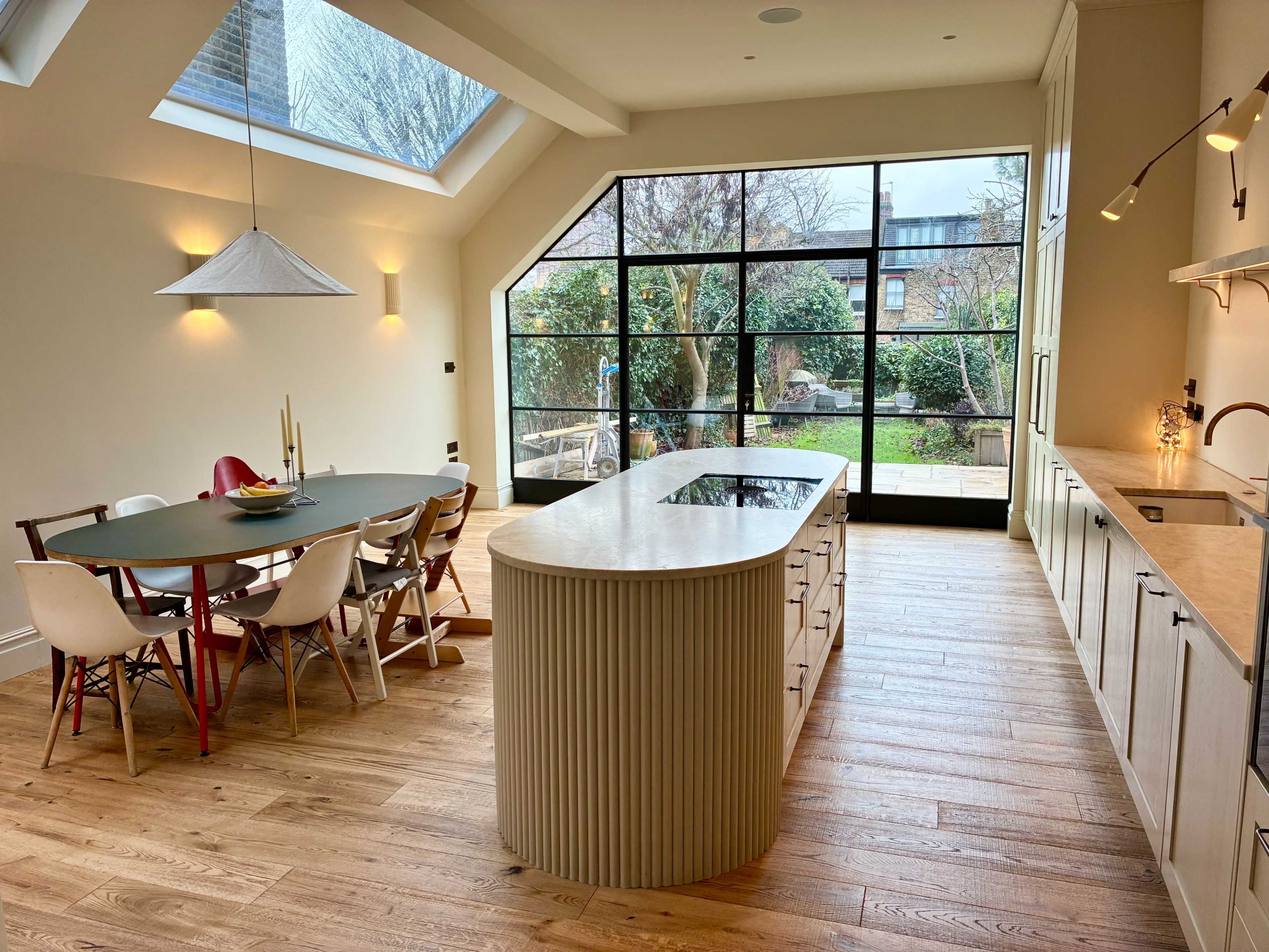 The image shows a modern kitchen and dining area with a large window overlooking a garden, featuring a round dining table and an island countertop.