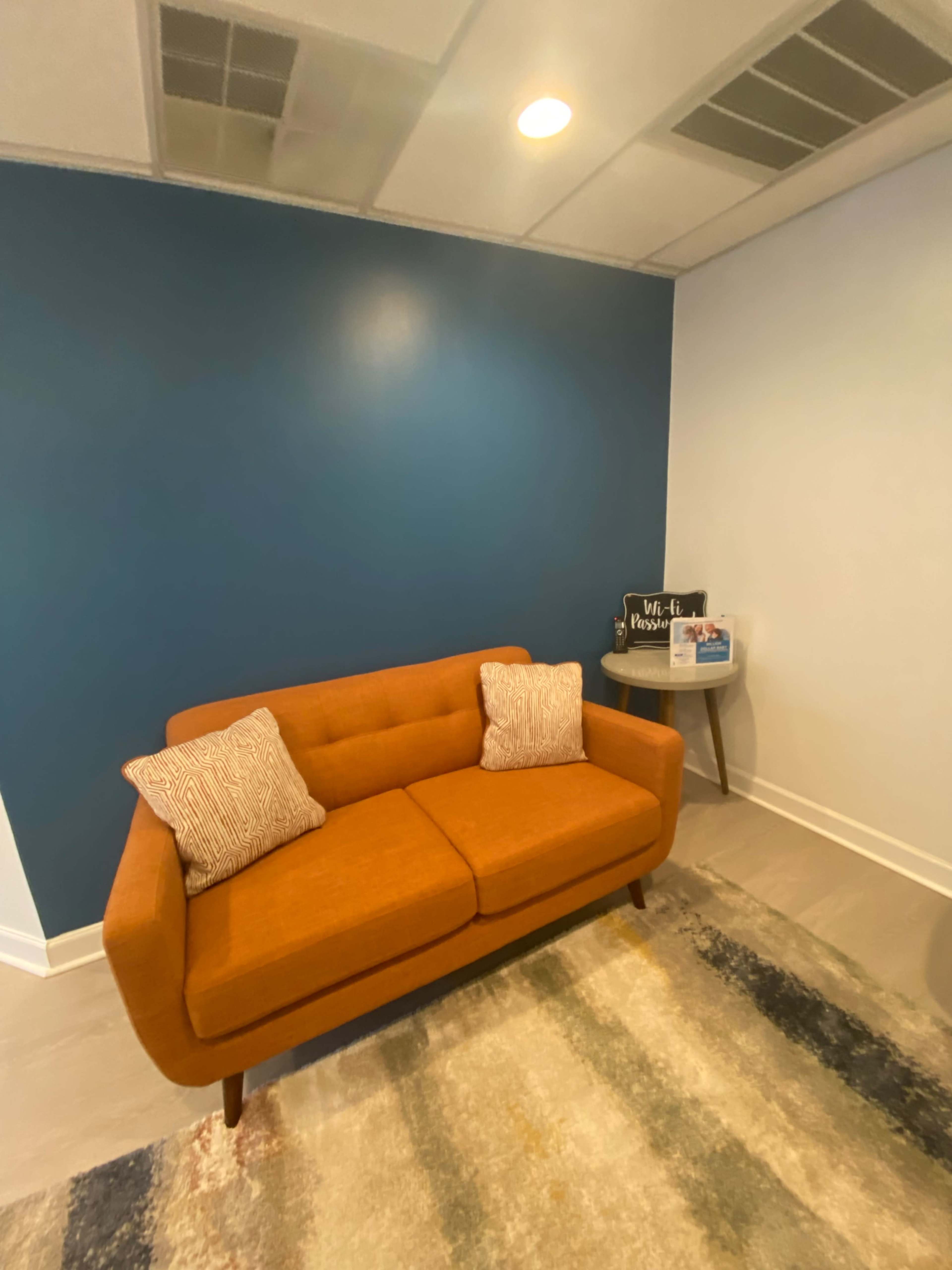 The image depicts an orange couch with two pillows against a blue wall in a room with light-colored flooring and a small side table.