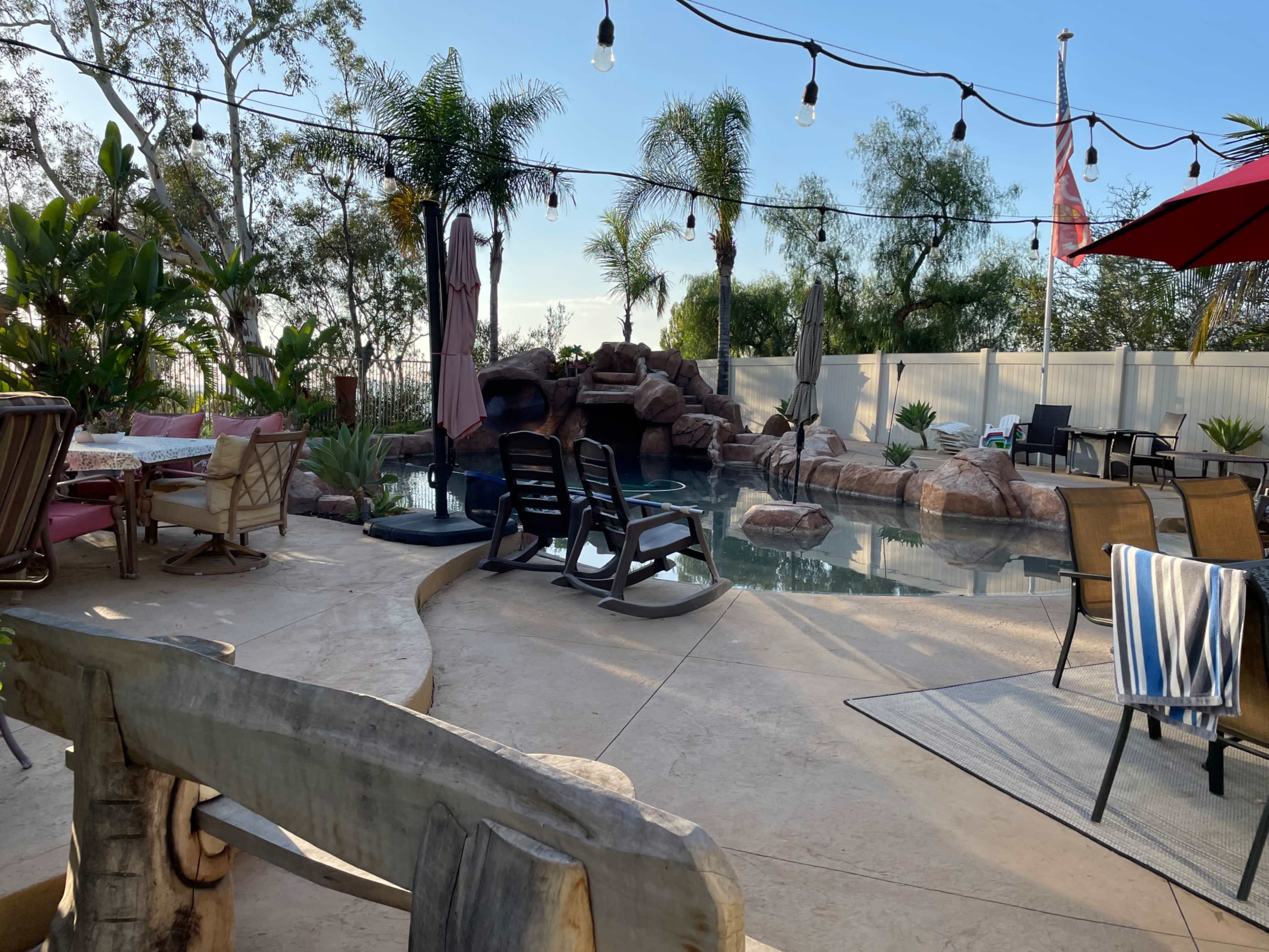 A backyard pool area featuring lounge chairs, a rock waterfall, palm trees, and string lights overhead.