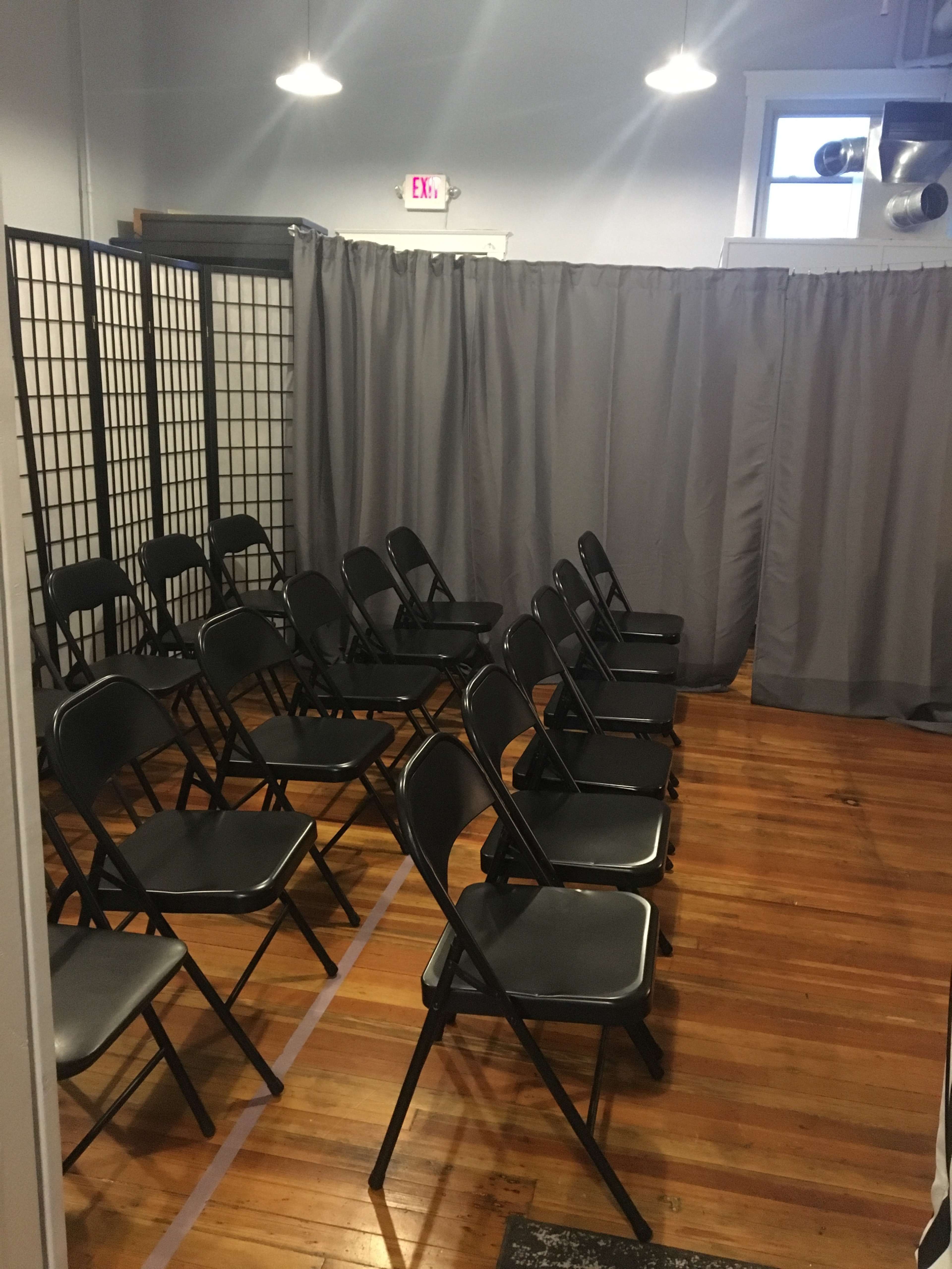 A room with several rows of black folding chairs set up in front of a gray curtain and partition screen.