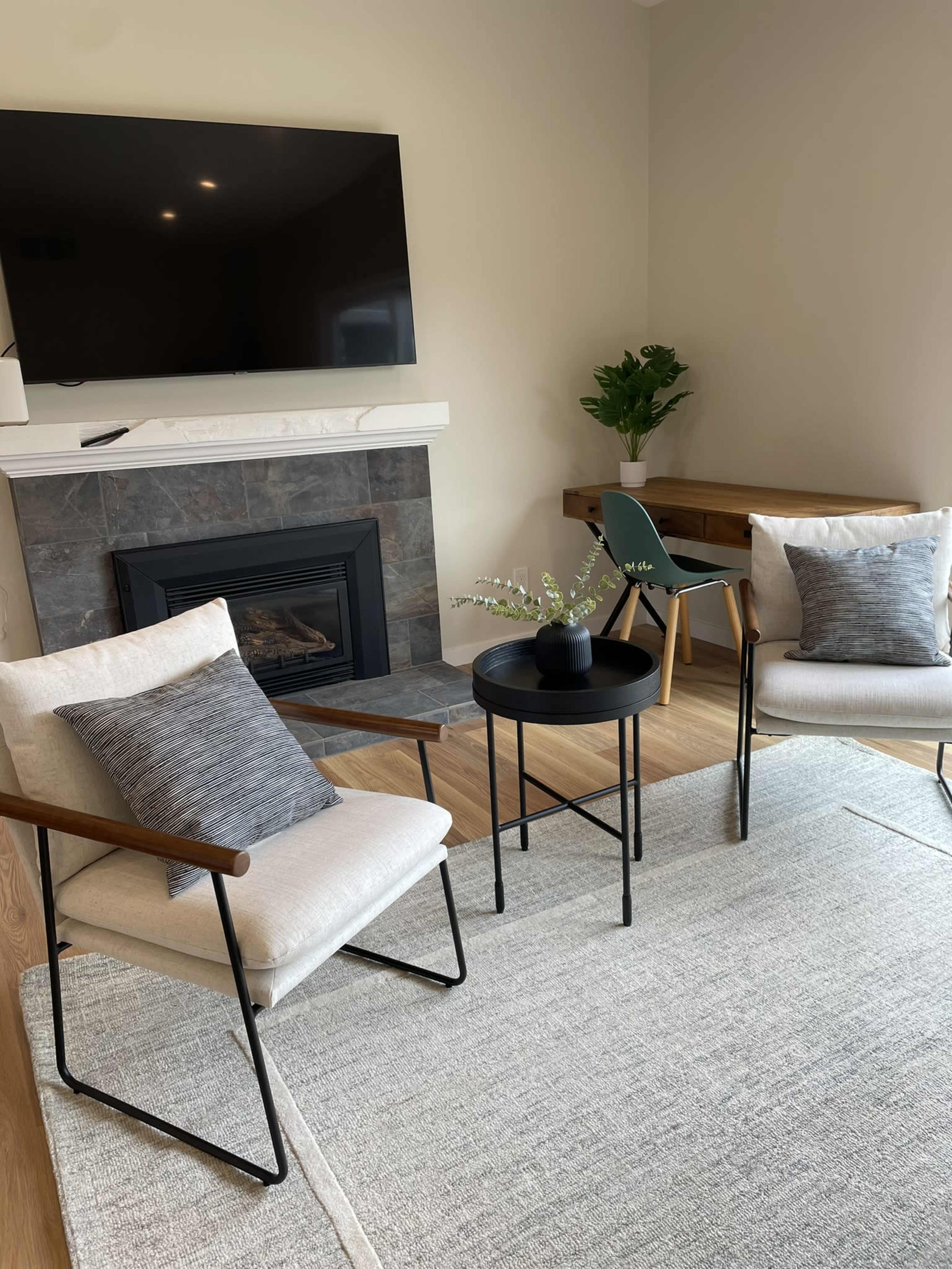The image shows a living space with two upholstered chairs, a round black table, a fireplace, a television mounted on the wall, and a small dining table with a green chair.