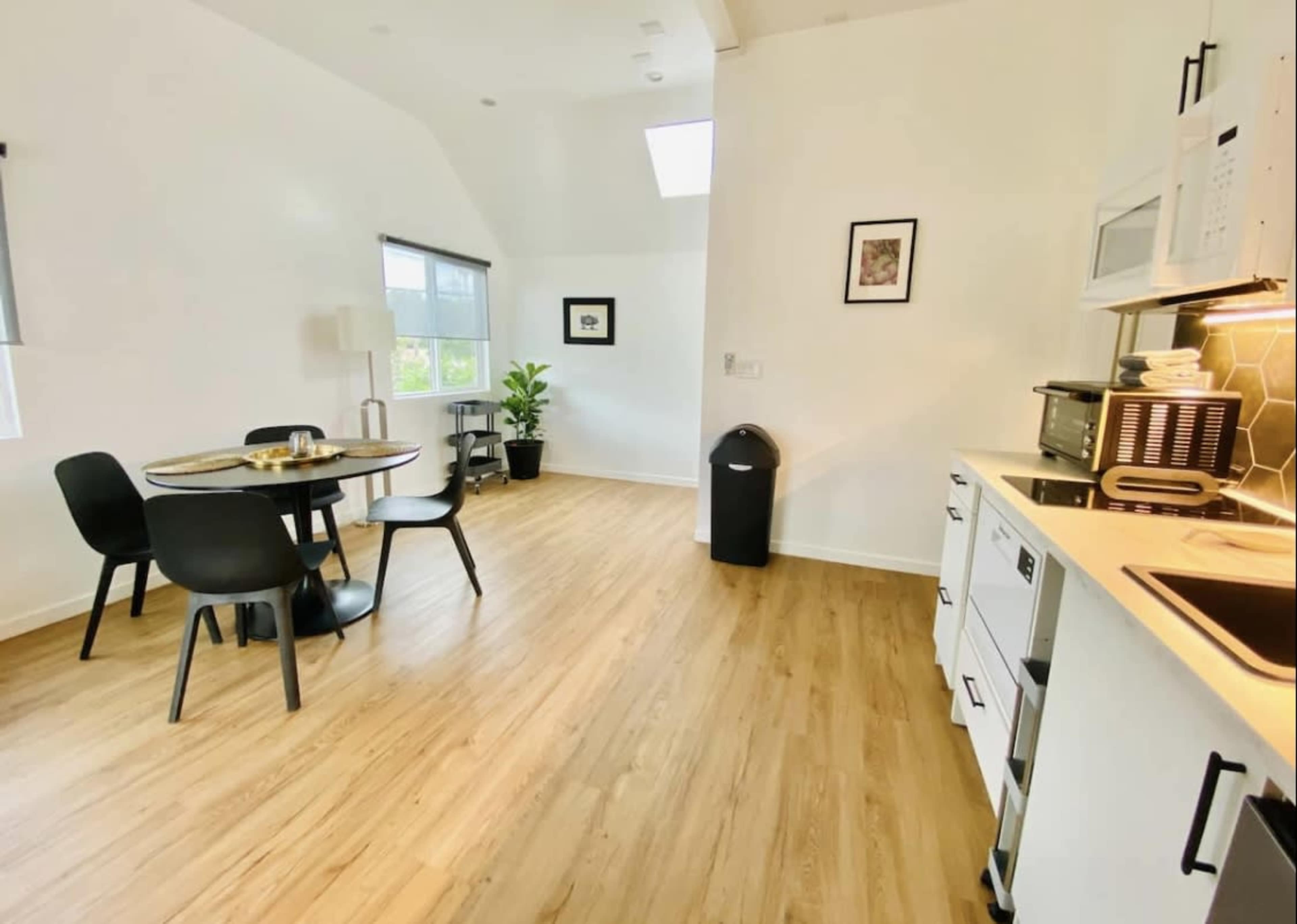 A modern kitchen and dining area feature a round table with four black chairs, white cabinetry, and a large window that allows natural light to flood the space.