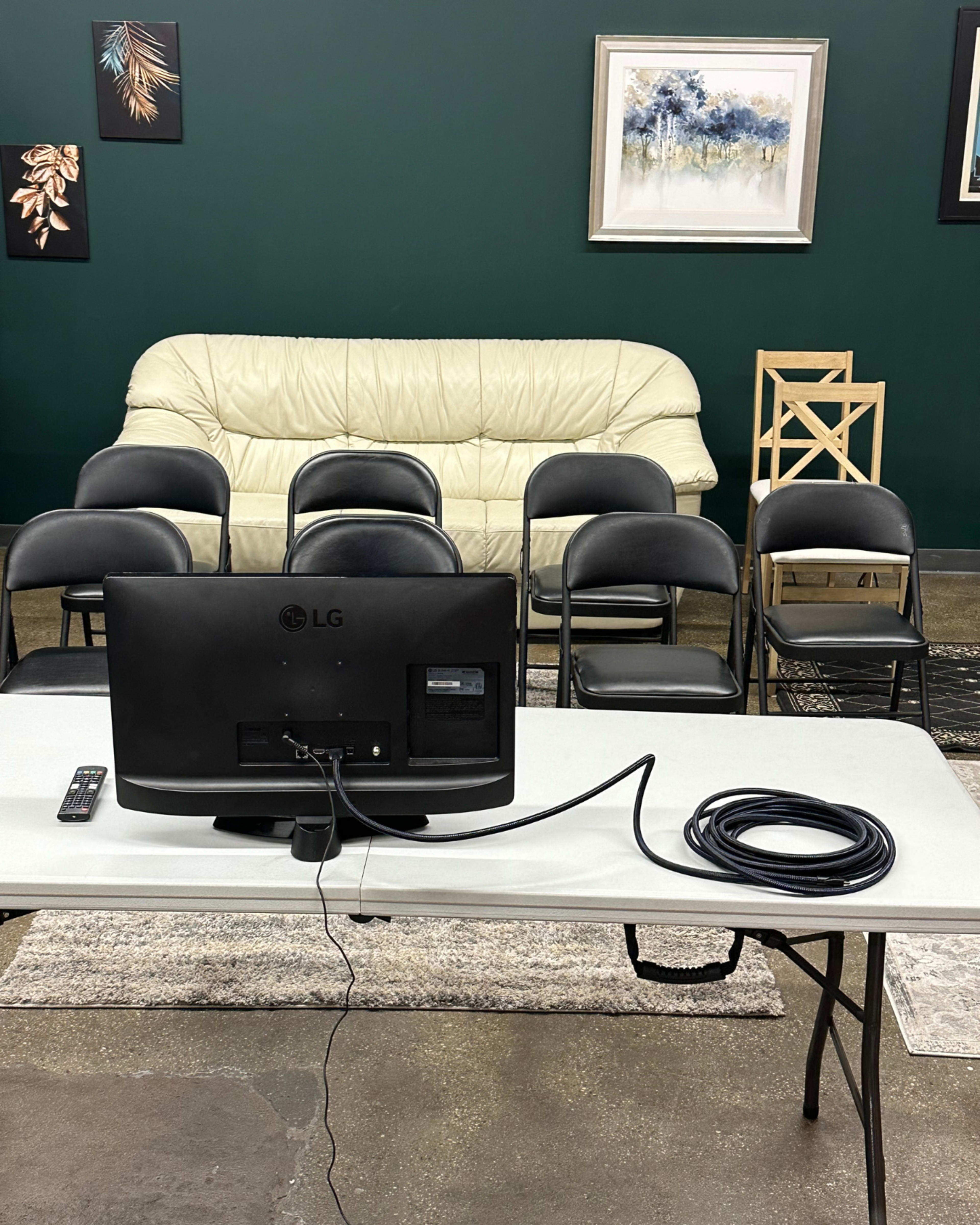 A white sofa is positioned against a green wall, facing a table with an LG monitor and several black folding chairs arranged in front of it.