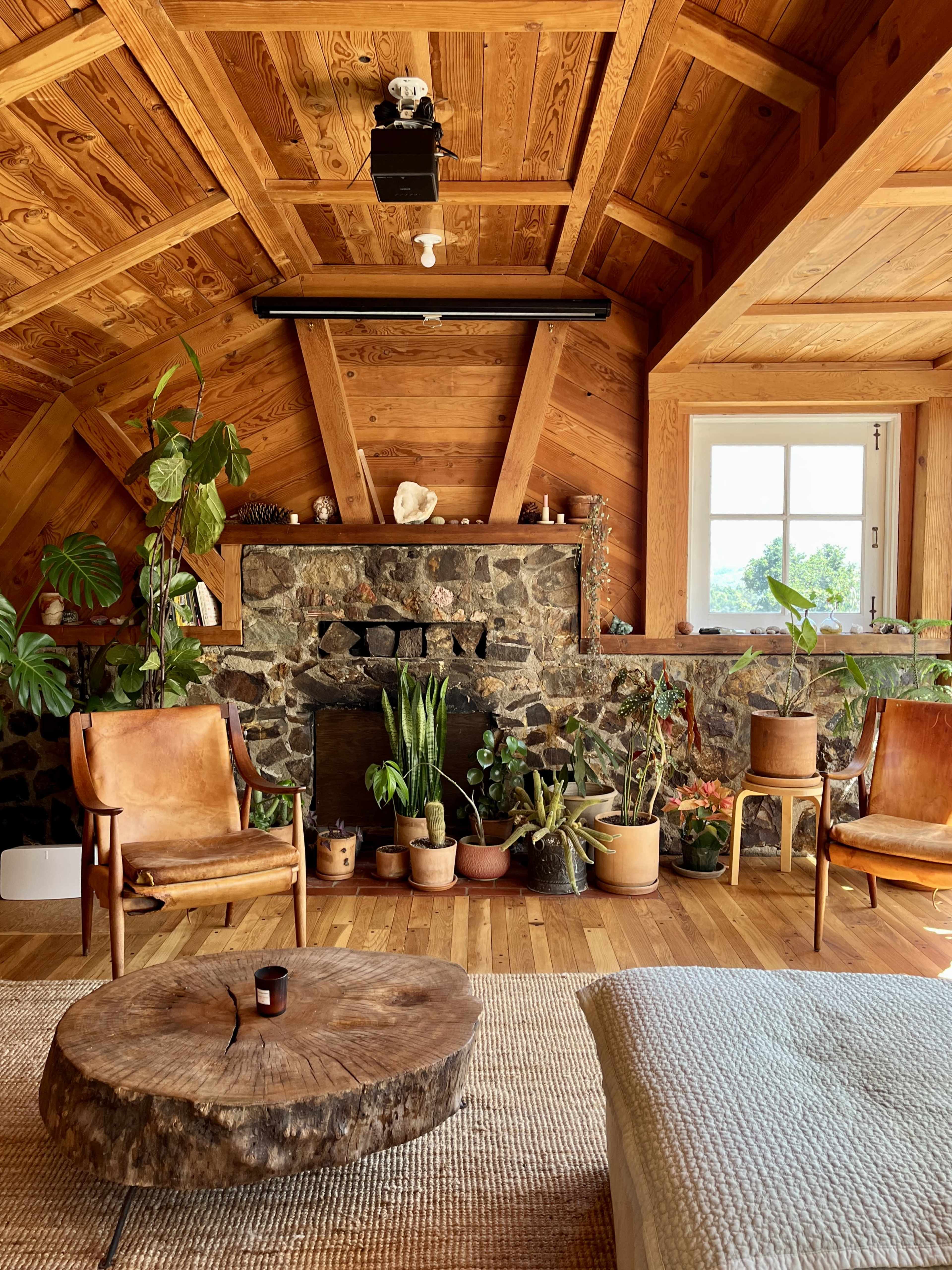 The interior of a wooden cabin features a stone fireplace, potted plants, two leather chairs, and a round wood coffee table.