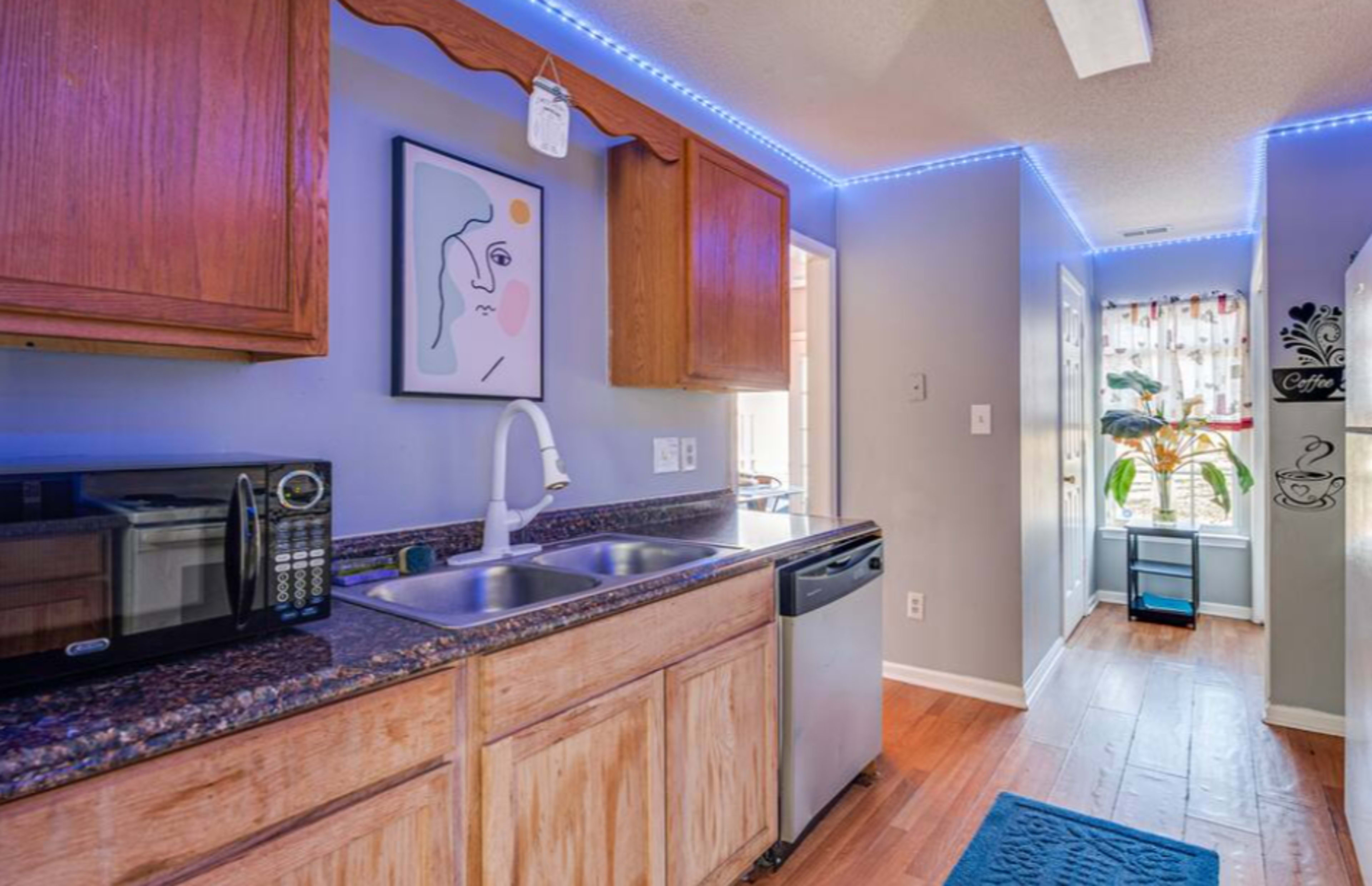 The image shows a kitchen with wooden cabinets, a double sink, a microwave, and a dishwasher, illuminated by blue LED strip lights along the ceiling.