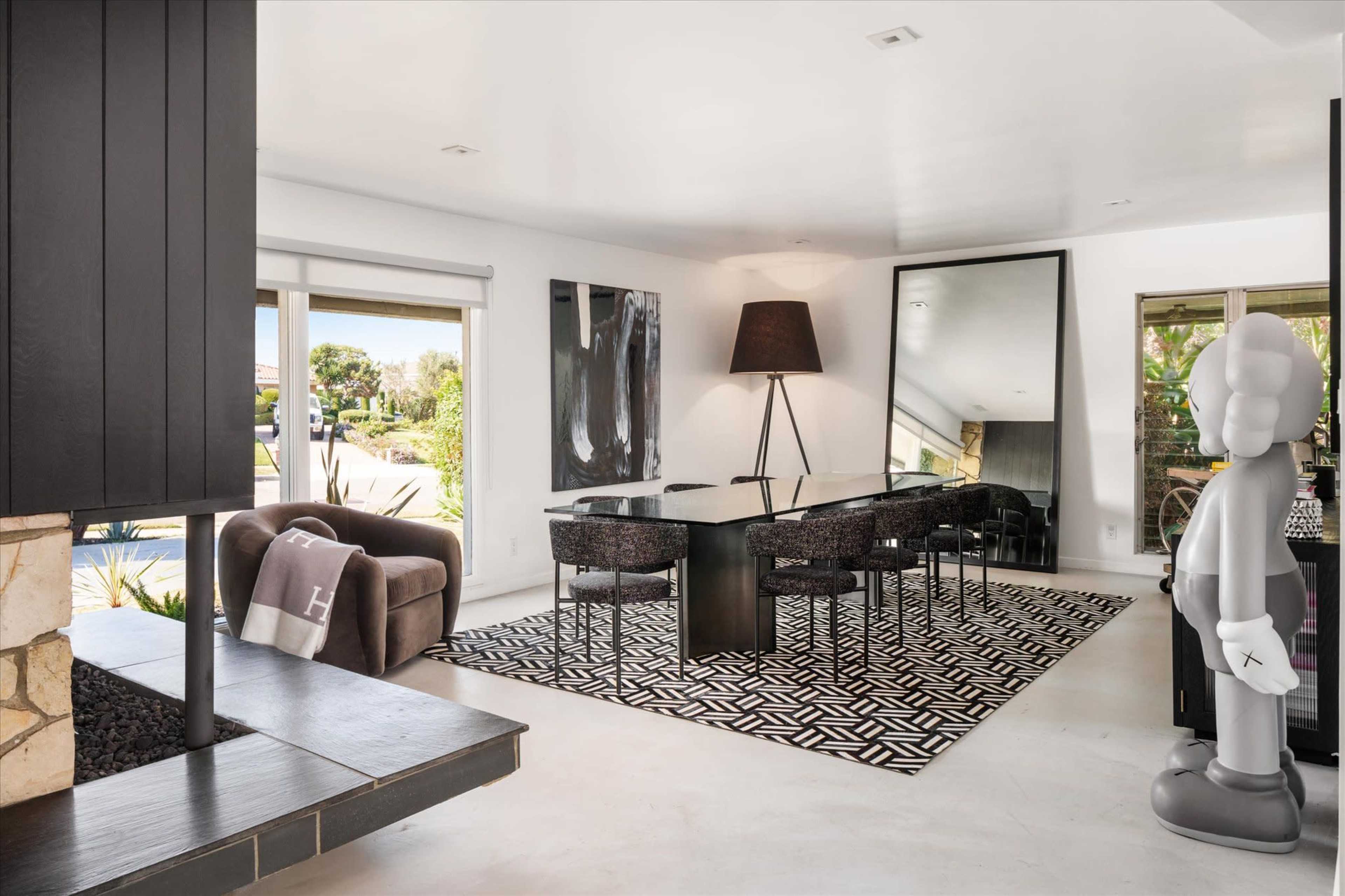 The image shows a modern dining area with a black dining table surrounded by dark chairs, a floor lamp, and a large mirror, all set against a backdrop of large windows letting in natural light.
